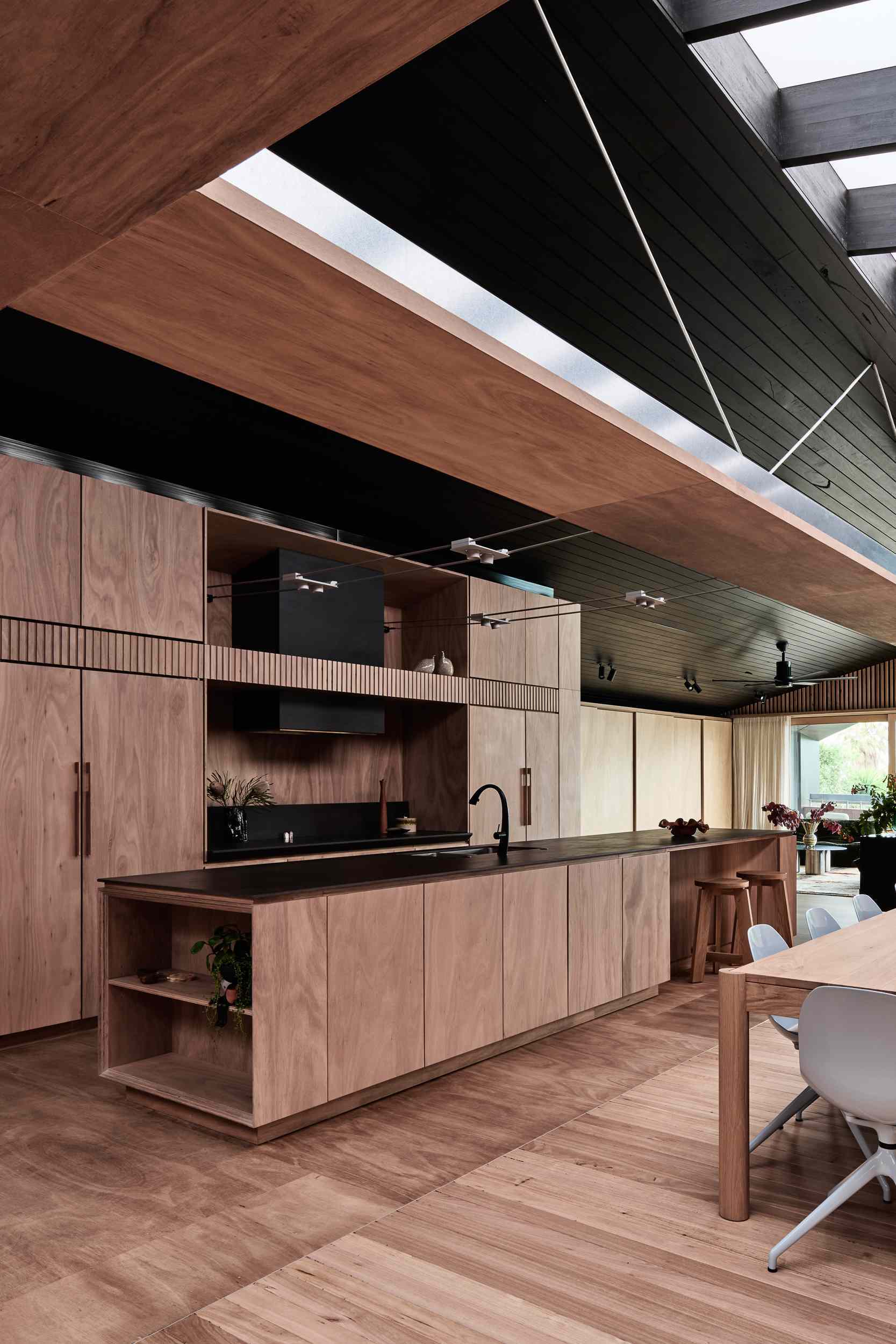 The kitchen combines natural timber and matte black accents for a warm, modern aesthetic. Seamless cabinetry and a dark porcelain countertop keep things clean and cohesive, while open shelving, a casual island bar, and exposed track lighting add laid-back, functional charm.