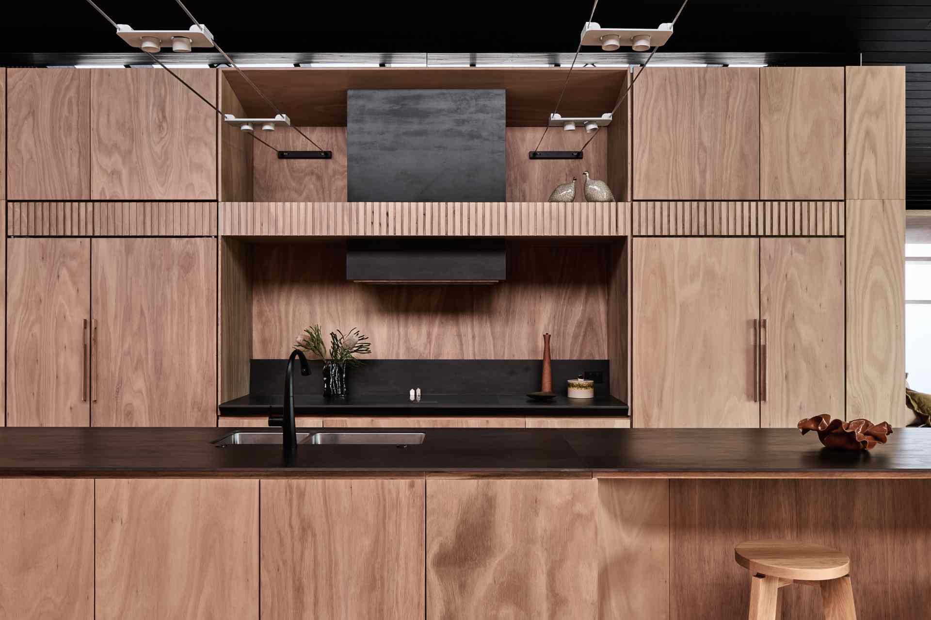 The kitchen combines natural timber and matte black accents for a warm, modern aesthetic. Seamless cabinetry and a dark porcelain countertop keep things clean and cohesive, while open shelving, a casual island bar, and exposed track lighting add laid-back, functional charm.