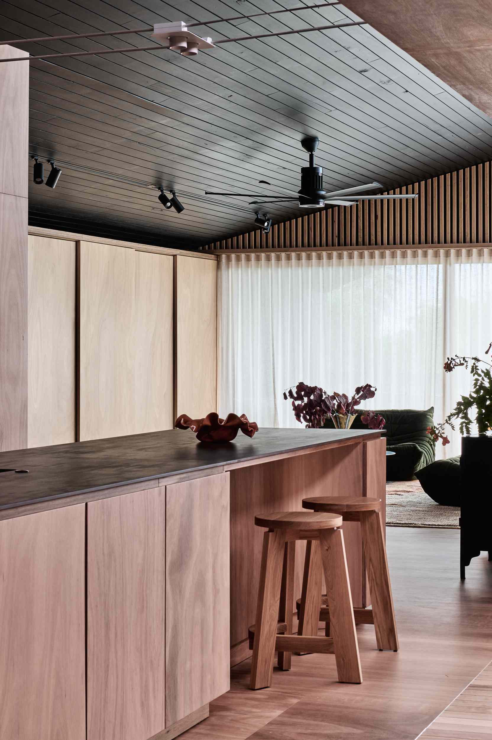 The kitchen combines natural timber and matte black accents for a warm, modern aesthetic. Seamless cabinetry and a dark porcelain countertop keep things clean and cohesive, while open shelving, a casual island bar, and exposed track lighting add laid-back, functional charm.