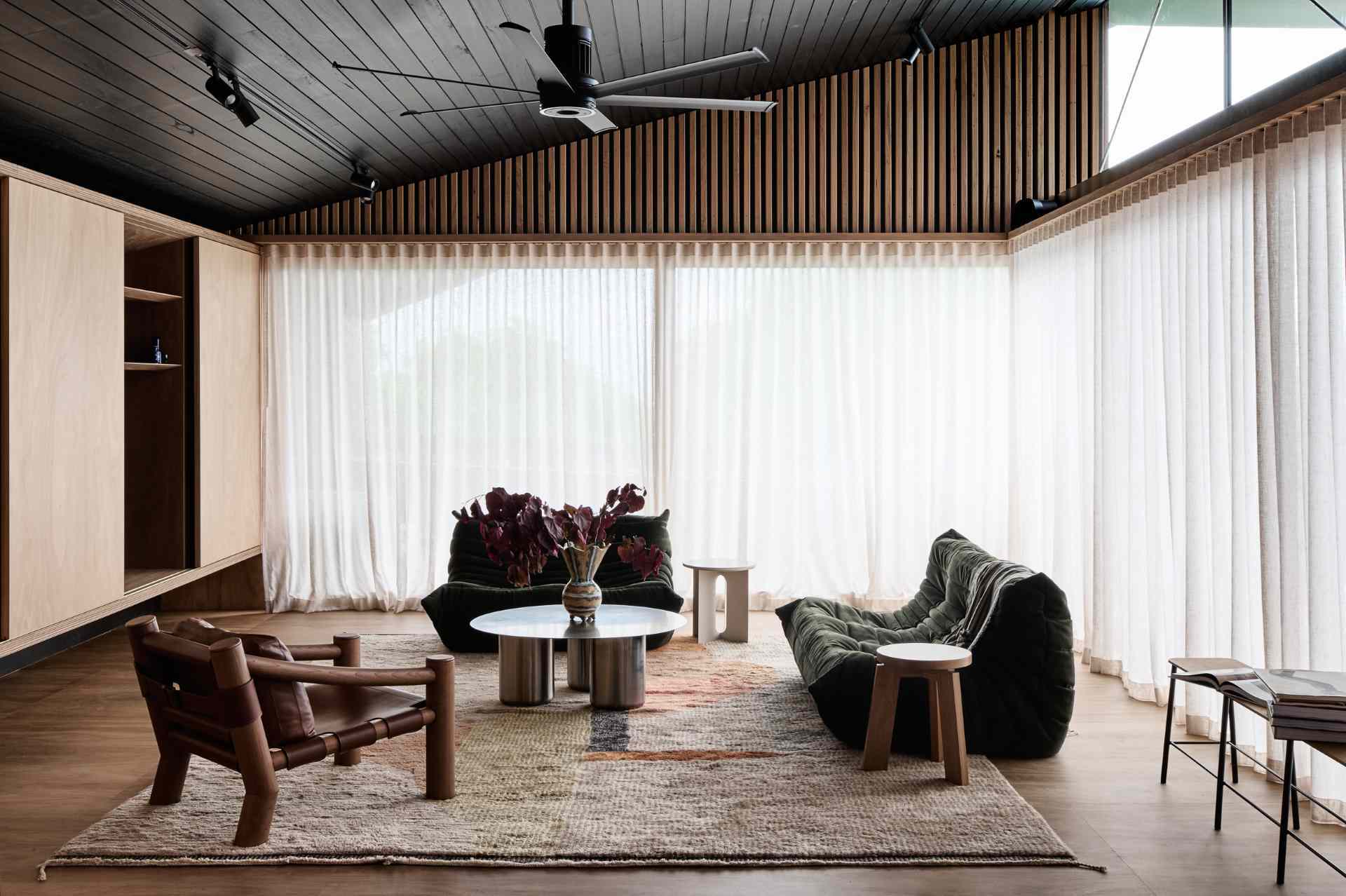 The living room exudes relaxed comfort with low-slung seating and deep olive tones that ground the light timber palette. Sculptural chairs and textured accents add visual interest, while sheer curtains gently filter light, softening the dark timber ceiling and warming the wood-lined space.