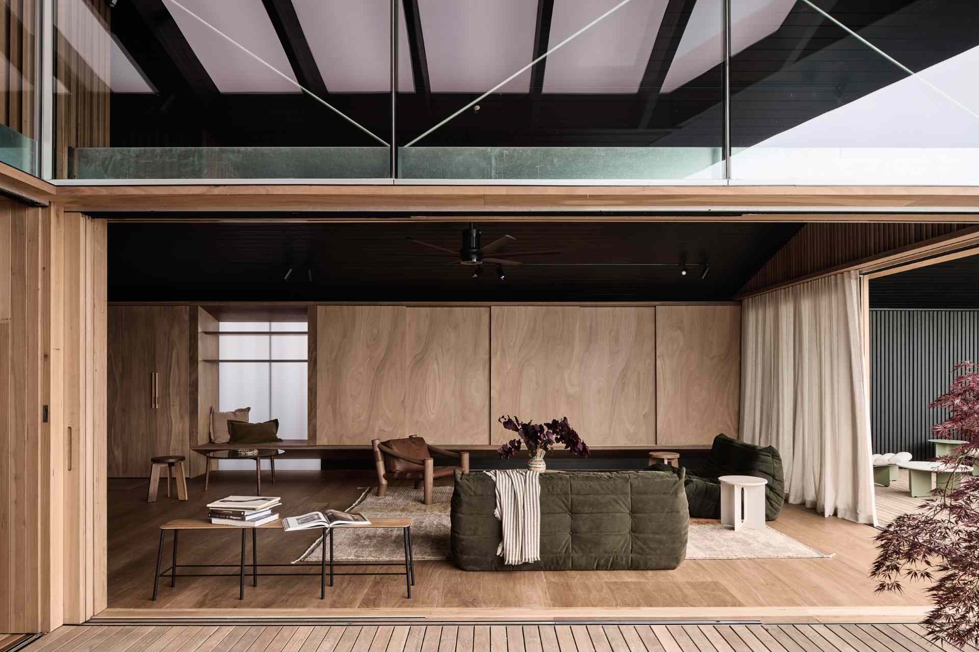 The living room exudes relaxed comfort with low-slung seating and deep olive tones that ground the light timber palette. Sculptural chairs and textured accents add visual interest, while sheer curtains gently filter light, softening the dark timber ceiling and warming the wood-lined space.