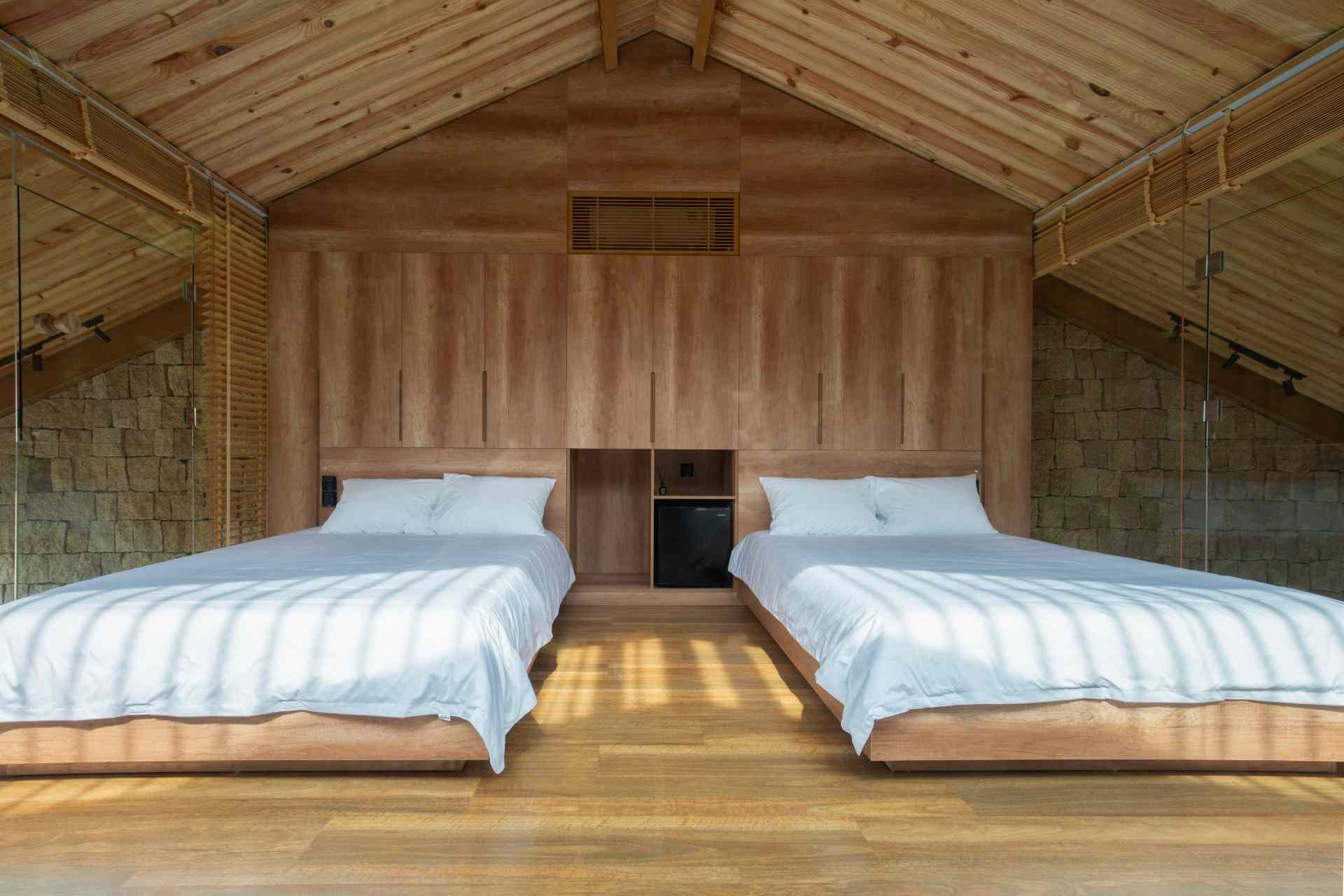 Bedroom with a pitched wooden ceiling, custom built-in cabinetry, and beds positioned between two walls of floor-to-ceiling glass that flood the space with natural light.