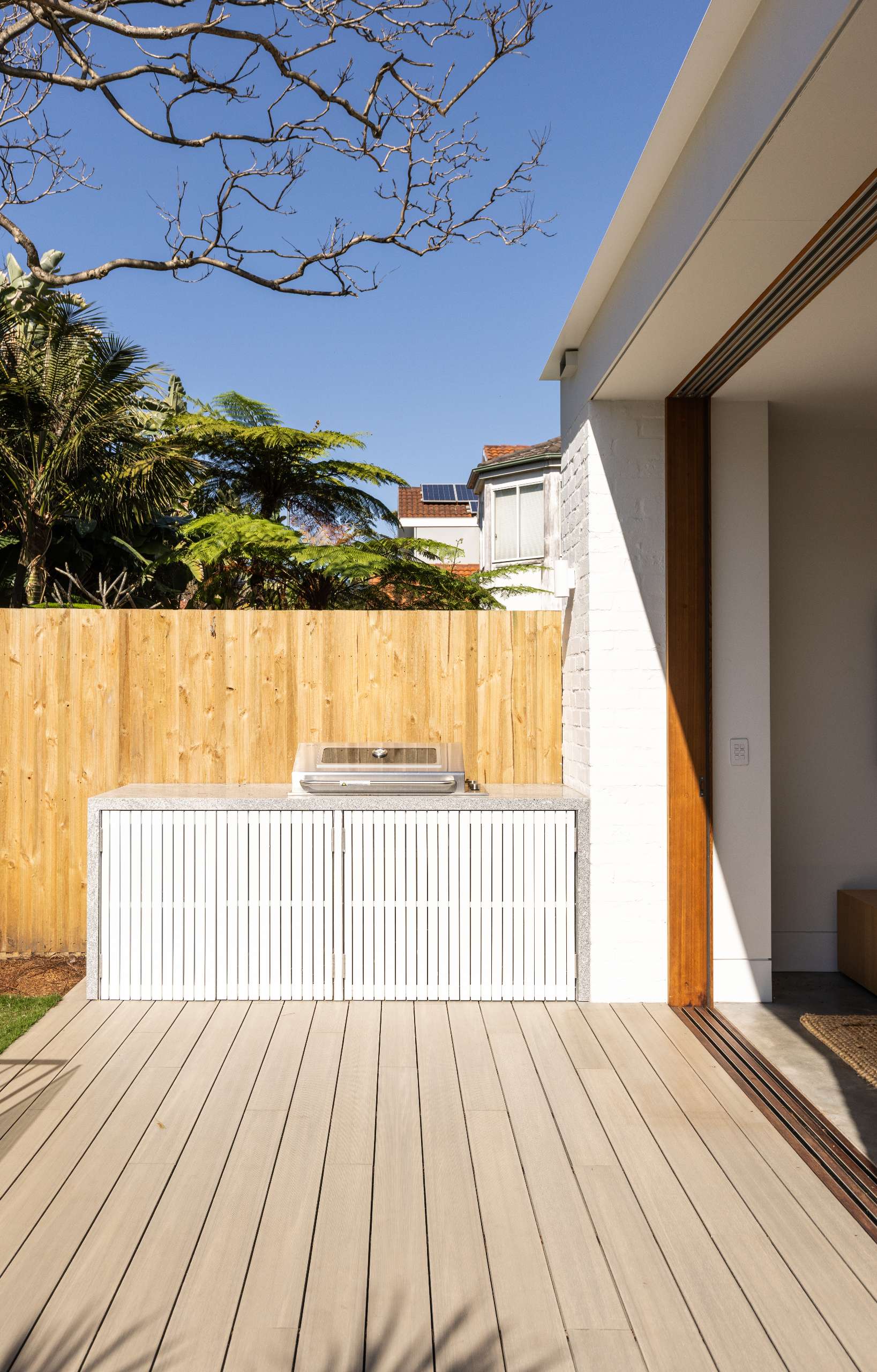 A sleek outdoor BBQ kitchen sits against a timber fence, featuring clean white battens with hidden storage, seamlessly extending from an open-plan living area onto a light timber deck.