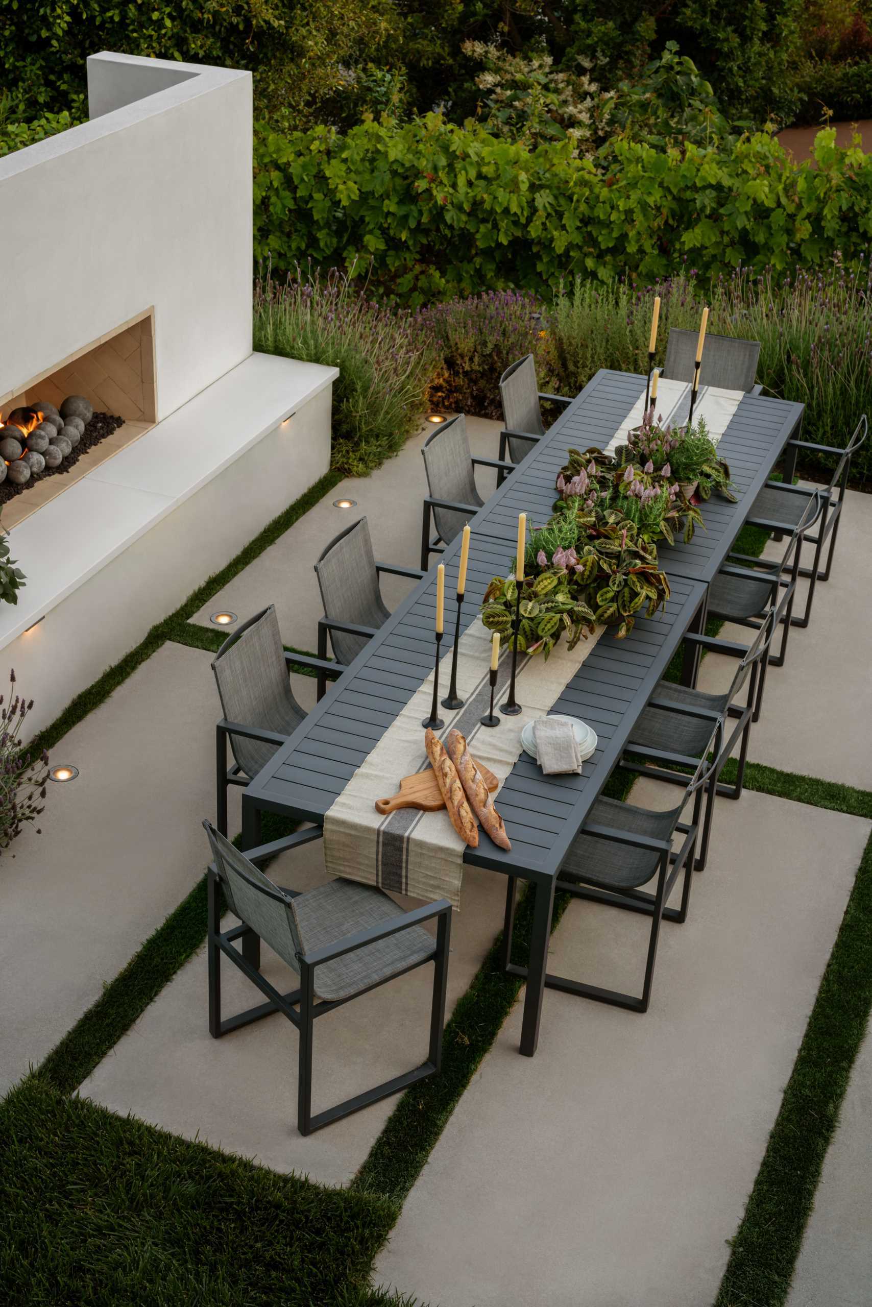 This contemporary outdoor dining space features a striking rectangular table with minimalist chairs, creating a clean and inviting setting for alfresco dining. Surrounded by carefully arranged greenery, including lavender, and clean stone pavers, the design emphasizes simplicity and elegance. Subtle lighting and a fireplace enhances the space, making it ideal for both daytime meals and evening gatherings, blending modern aesthetics with nature’s tranquility.