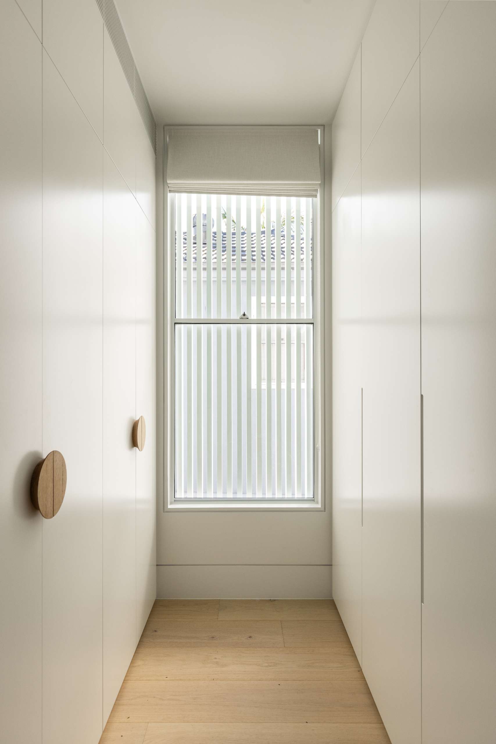 This walk-in wardrobe features full-height cabinetry with timber disc handles and soft battens that filter light through the window.
