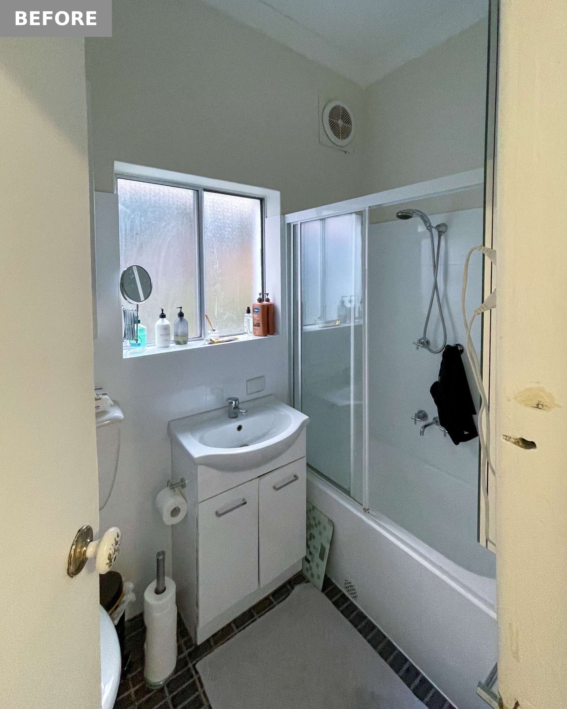 This original bathroom shows a compact, dated space with dark tile flooring, a basic vanity, and a combined shower-over-bath setup. Frosted glass windows provide privacy but limit natural light, while cluttered surfaces and aging fixtures contribute to the room’s cramped, utilitarian feel.