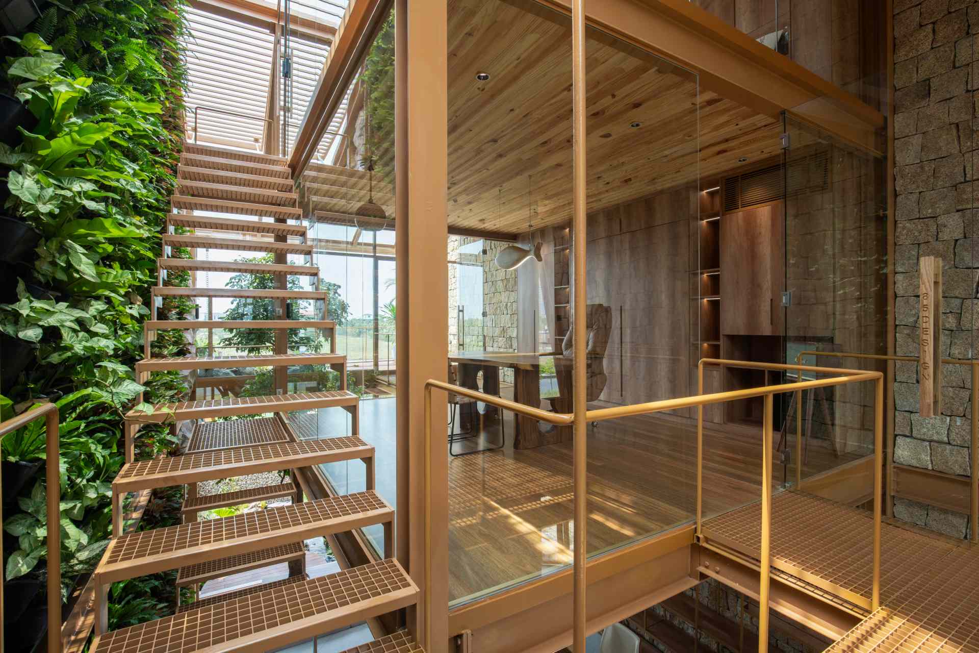 A light-filled home office enclosed in glass with views of a lush green wall. The wood-paneled wall behind the desk hides a fold-down bed, making it easy to switch from work mode to rest.