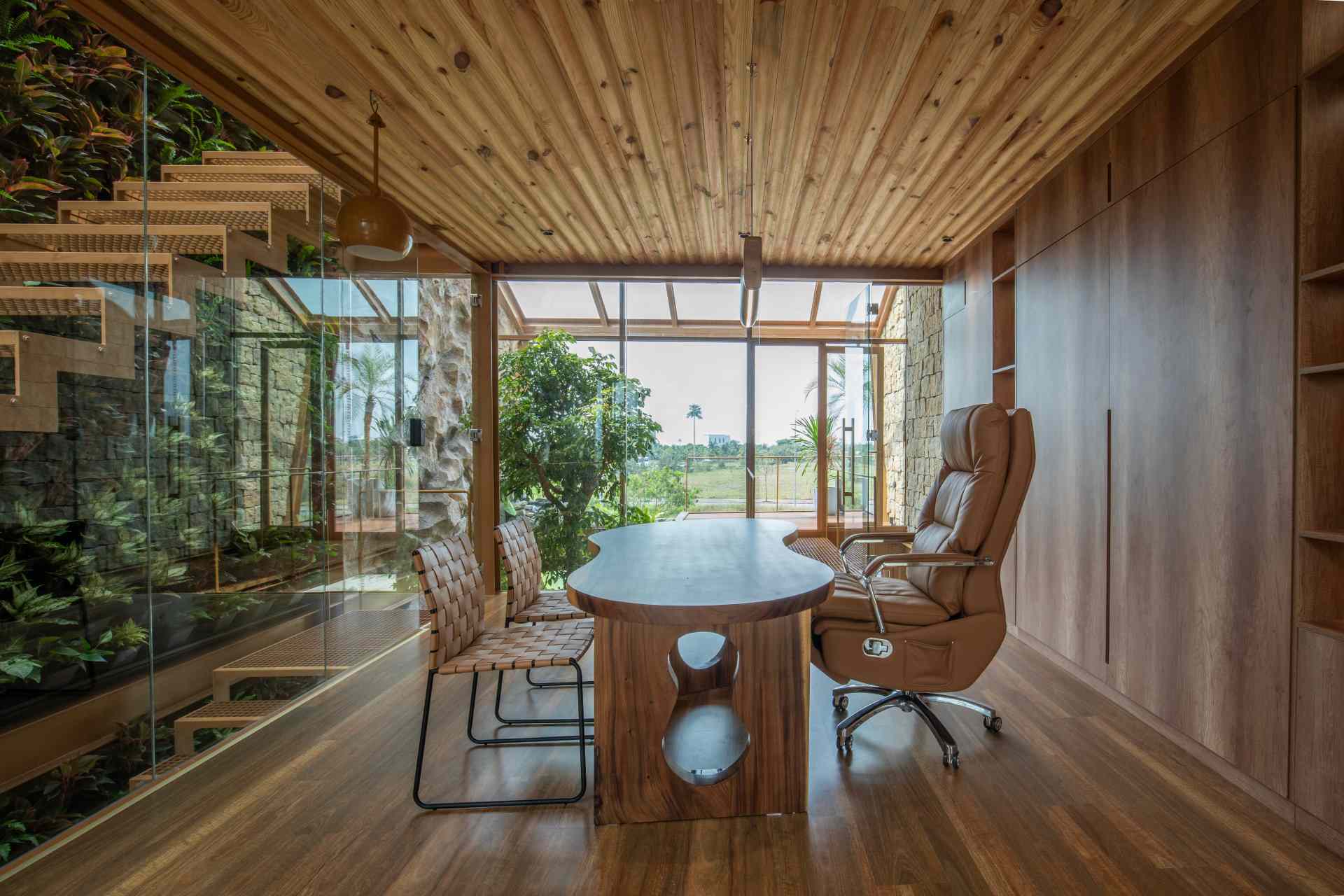 A light-filled home office enclosed in glass with views of a lush green wall. The wood-paneled wall behind the desk hides a fold-down bed, making it easy to switch from work mode to rest.