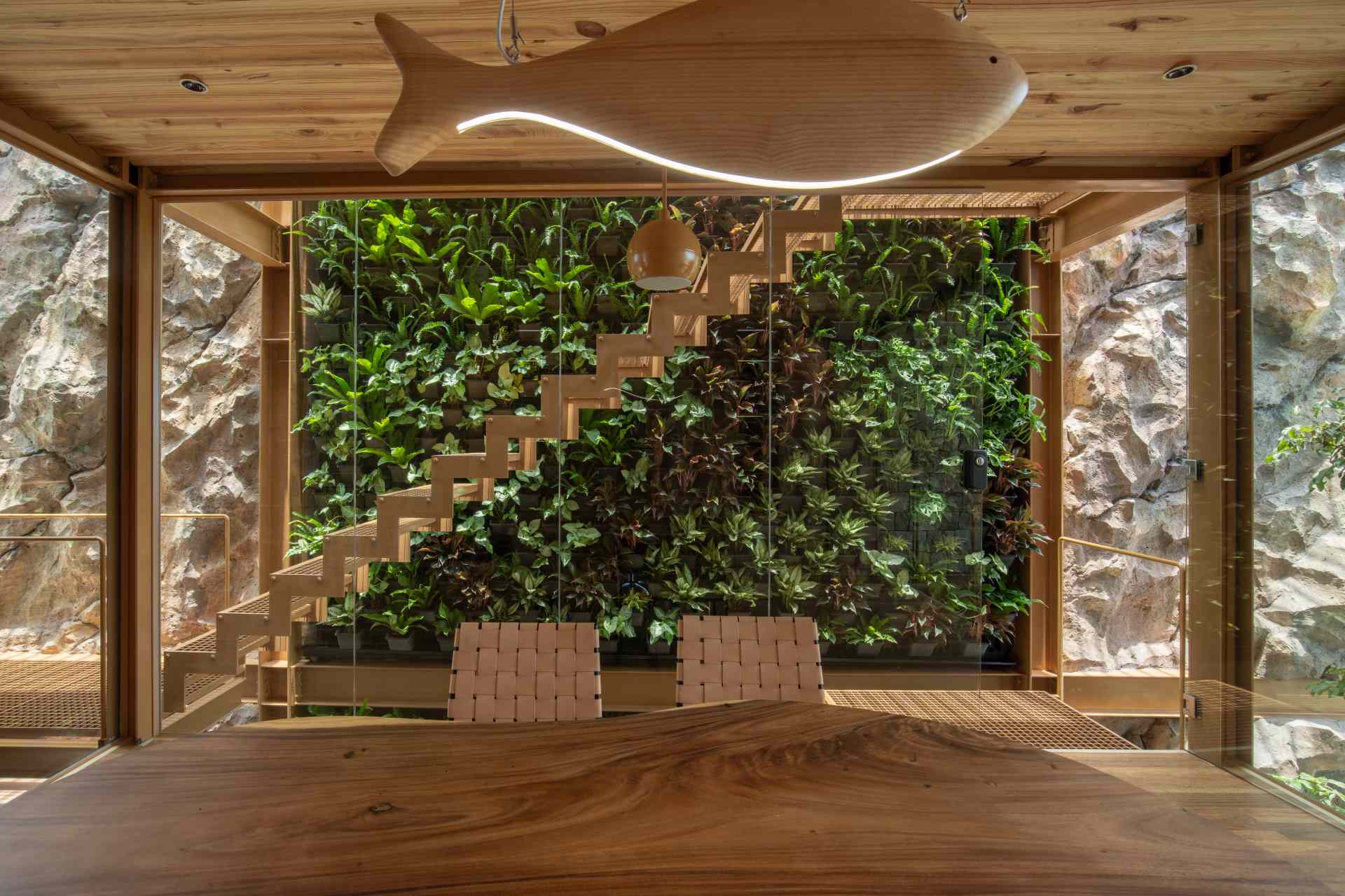 A light-filled home office enclosed in glass with views of a lush green wall. The wood-paneled wall behind the desk hides a fold-down bed, making it easy to switch from work mode to rest.