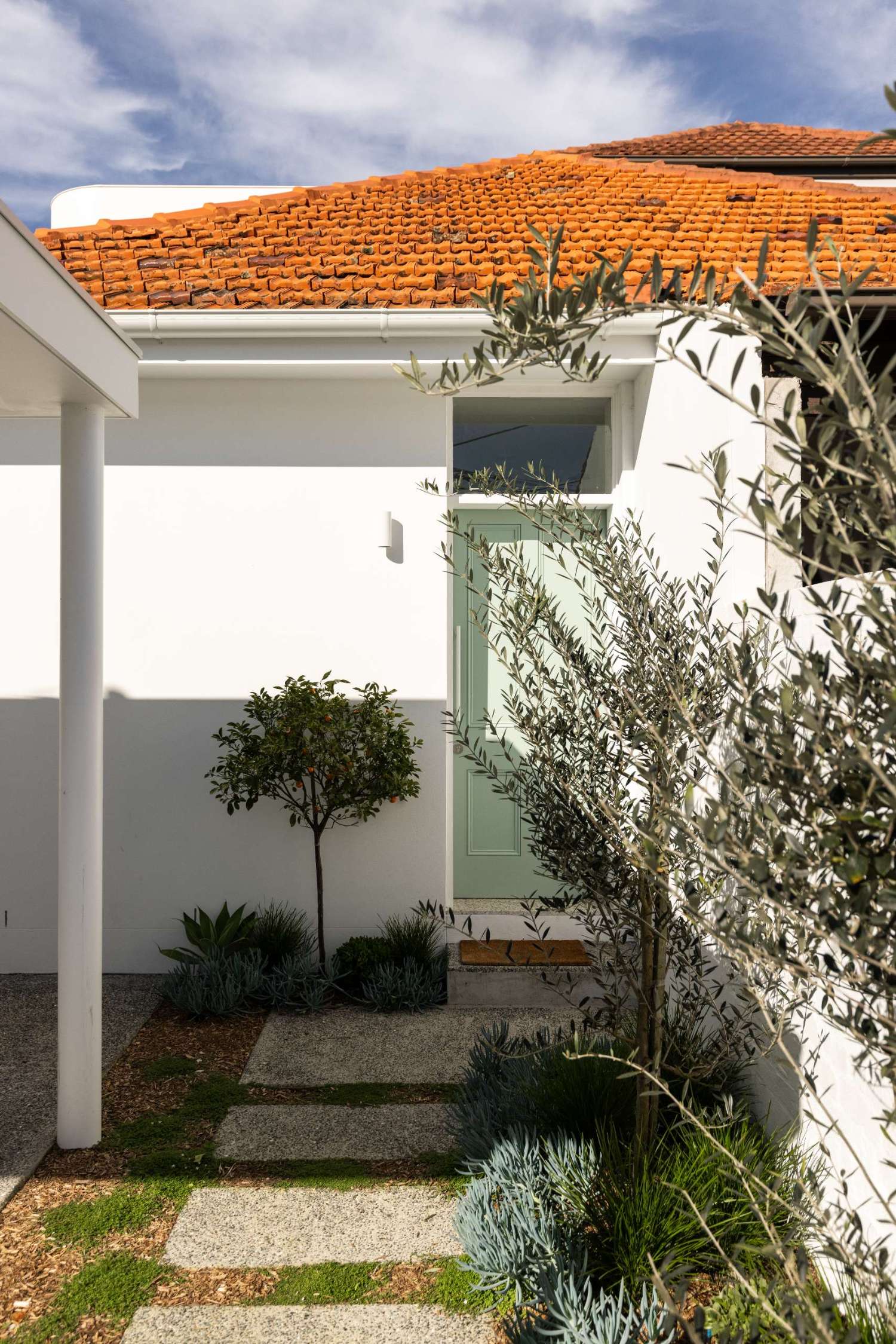 The original courtyard featured dated French doors, exposed utilities, and uneven paving. After the renovation, a sleek white facade, and fresh landscaping give the entry a serene, design-forward look. A pistachio green door complete the clean, coastal feel.