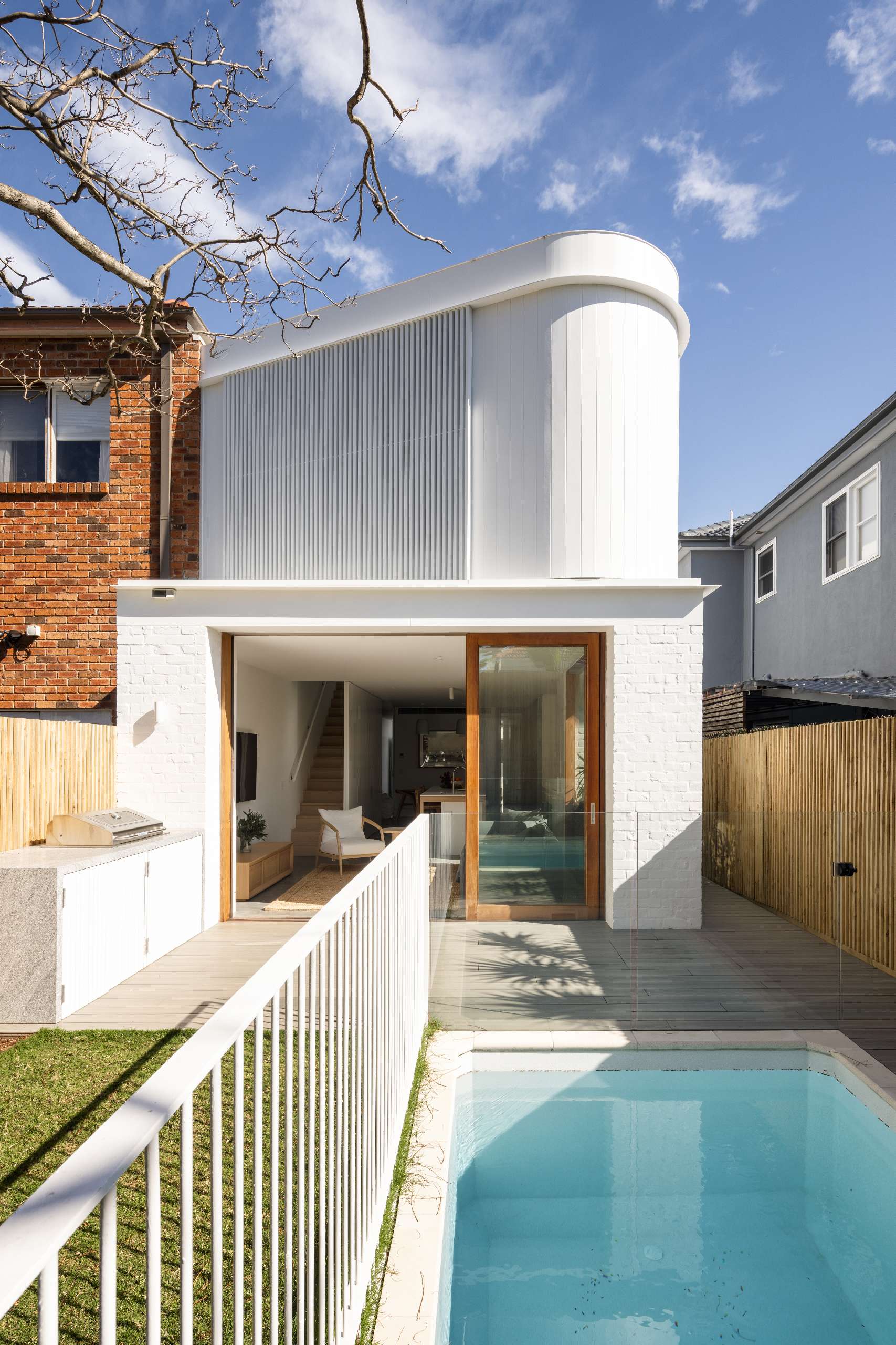 The once-boxy exterior was replaced with a two-level sculptural form wrapped in white render and curved vertical cladding. Timber-framed sliding glass doors invite seamless transition between inside and out, opening directly onto a new lawn, deck, and in-ground pool.