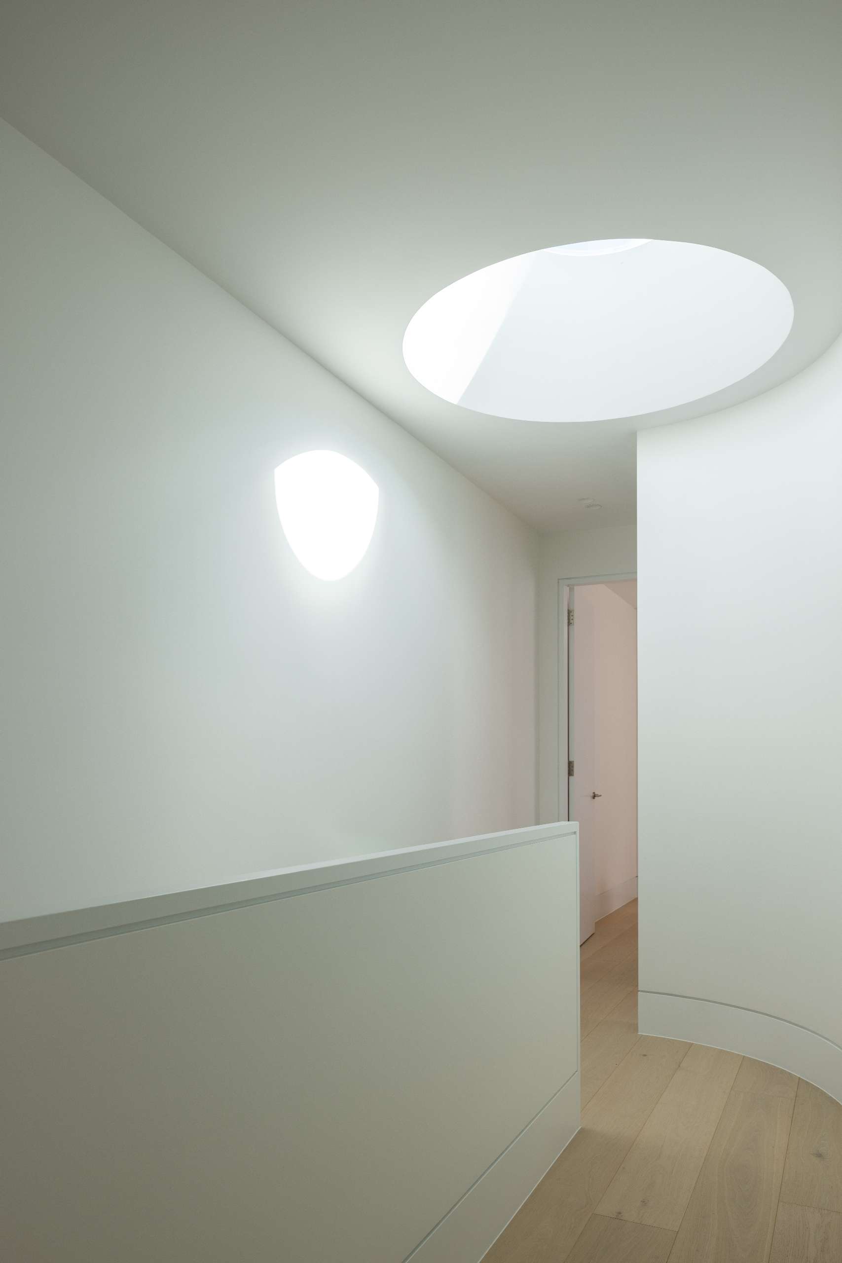 A circular skylight above the stairwell draws natural light into the heart of the home, creating a quiet moment of drama. This oculus not only improves passive solar performance but also casts shifting shadows that soften the minimalist palette and highlight the home's curved interior lines.