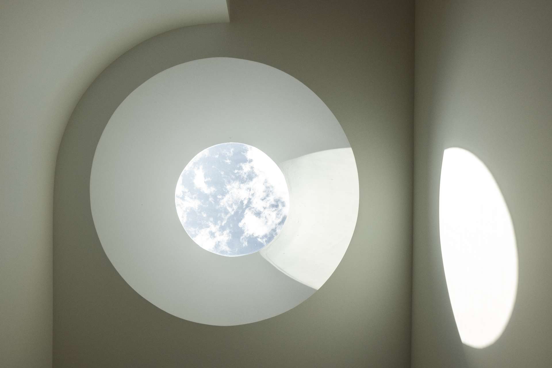 A circular skylight above the stairwell draws natural light into the heart of the home, creating a quiet moment of drama. This oculus not only improves passive solar performance but also casts shifting shadows that soften the minimalist palette and highlight the home's curved interior lines.