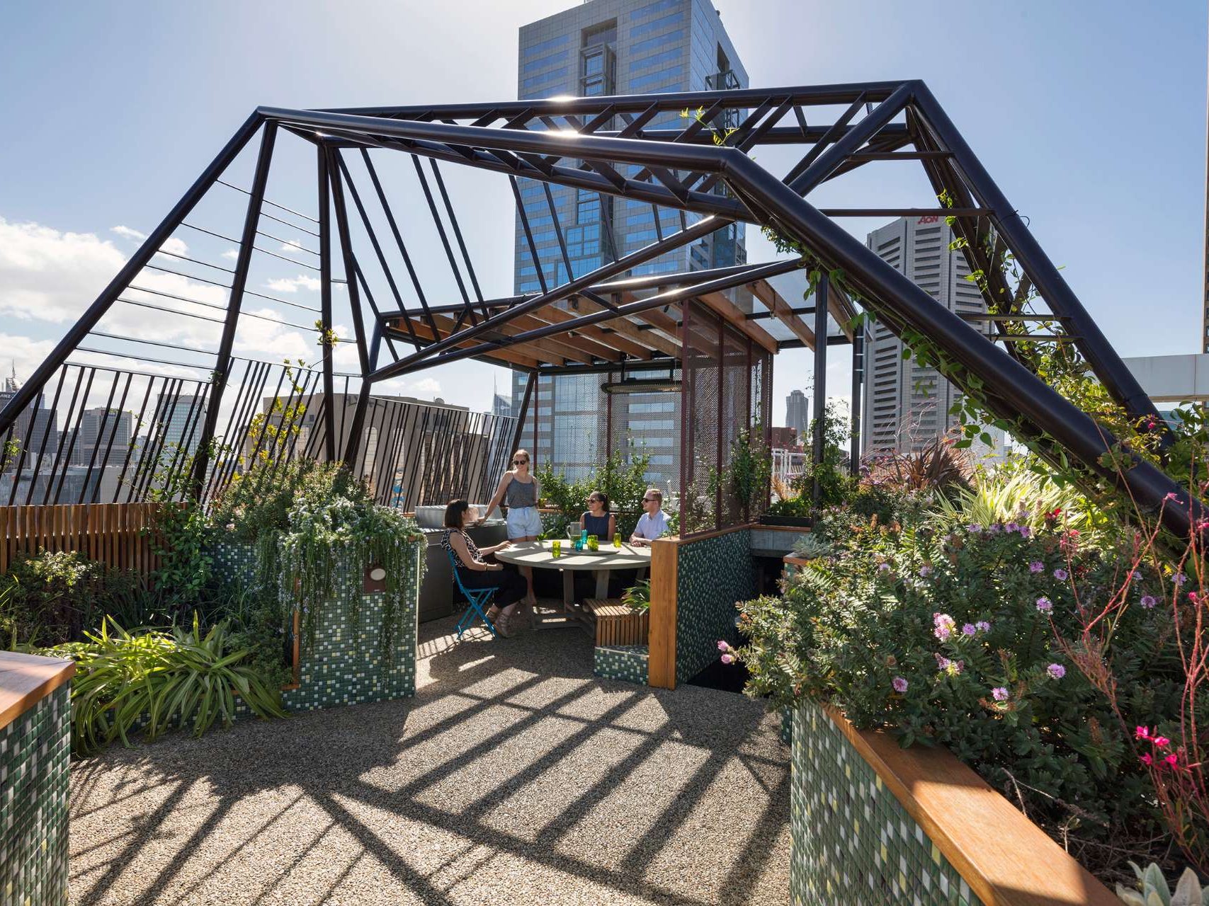 A sculptural steel arbour frames the Phoenix Rooftop garden in central Melbourne, casting dynamic shadows across a mosaic-tiled courtyard. The space blends lush planting, custom tiling, and defined seating zones to create a liveable garden 30 storeys above the city. This rooftop retreat shows how structure and greenery can work together in high-rise outdoor design.