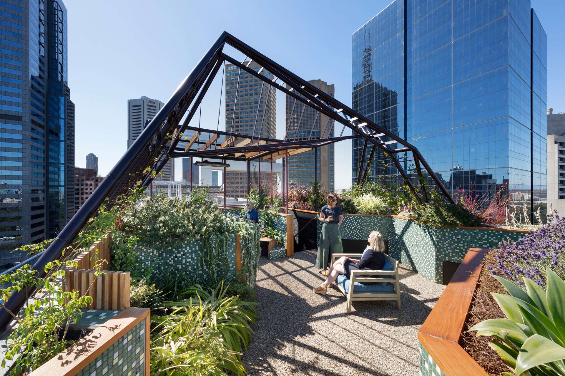 A sculptural steel arbour frames the Phoenix Rooftop garden in central Melbourne, casting dynamic shadows across a mosaic-tiled courtyard. The space blends lush planting, custom tiling, and defined seating zones to create a liveable garden 30 storeys above the city. This rooftop retreat shows how structure and greenery can work together in high-rise outdoor design.