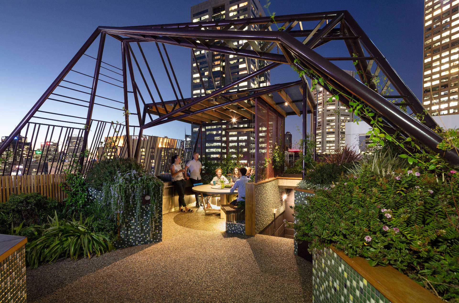 A sculptural steel arbour frames the Phoenix Rooftop garden in central Melbourne, casting dynamic shadows across a mosaic-tiled courtyard. The space blends lush planting, custom tiling, and defined seating zones to create a liveable garden 30 storeys above the city. This rooftop retreat shows how structure and greenery can work together in high-rise outdoor design.
