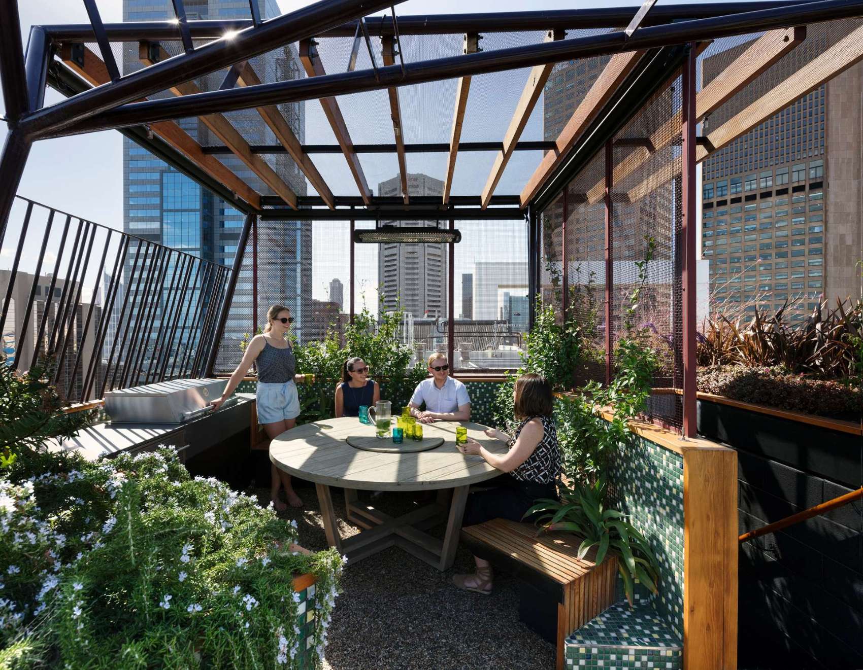 A sculptural steel arbour frames the Phoenix Rooftop garden in central Melbourne, casting dynamic shadows across a mosaic-tiled courtyard. A round table with nearby bbq creates an outdoor dining experience.