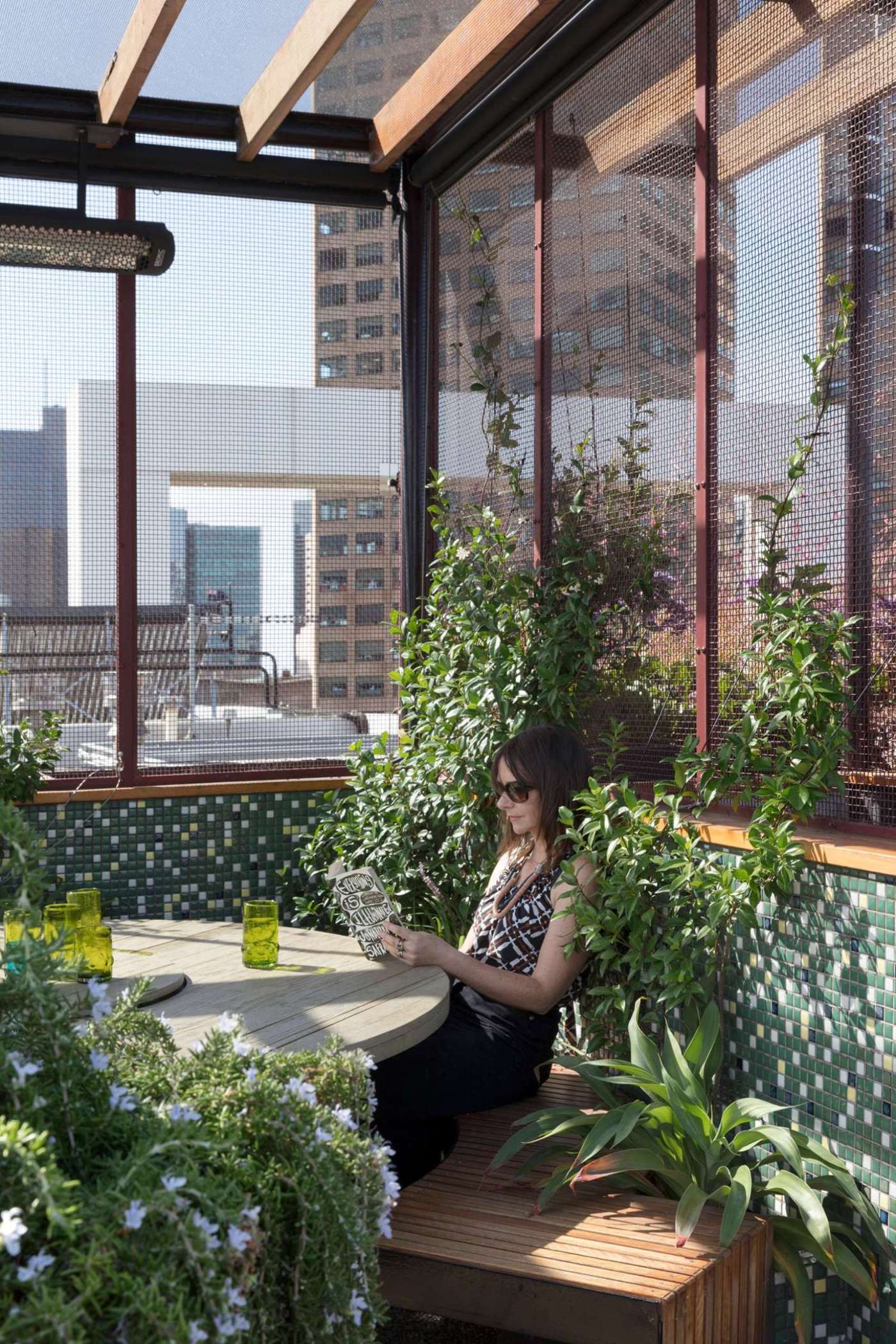 A sculptural steel arbour frames the Phoenix Rooftop garden in central Melbourne, casting dynamic shadows across a mosaic-tiled courtyard. A round table with nearby bbq creates an outdoor dining experience.