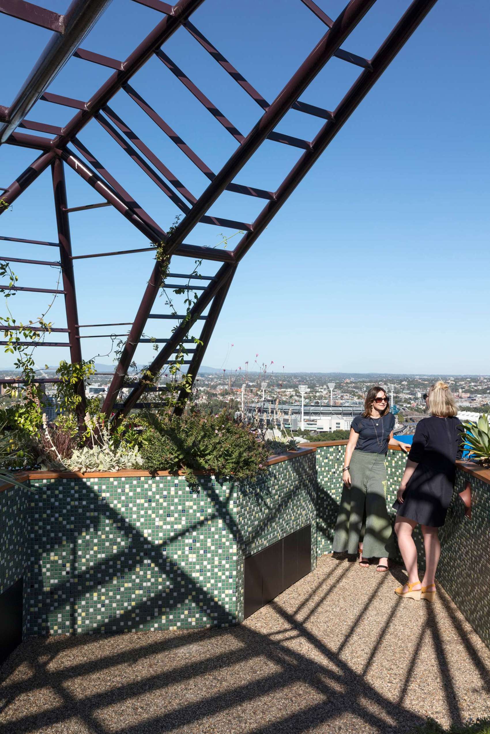 A rooftop planter filled with layered greenery creates a private pocket above the city, framed by a striking steel arbour and patterned mosaic tiles. A quiet corner with a view, perfect for conversation and connection.