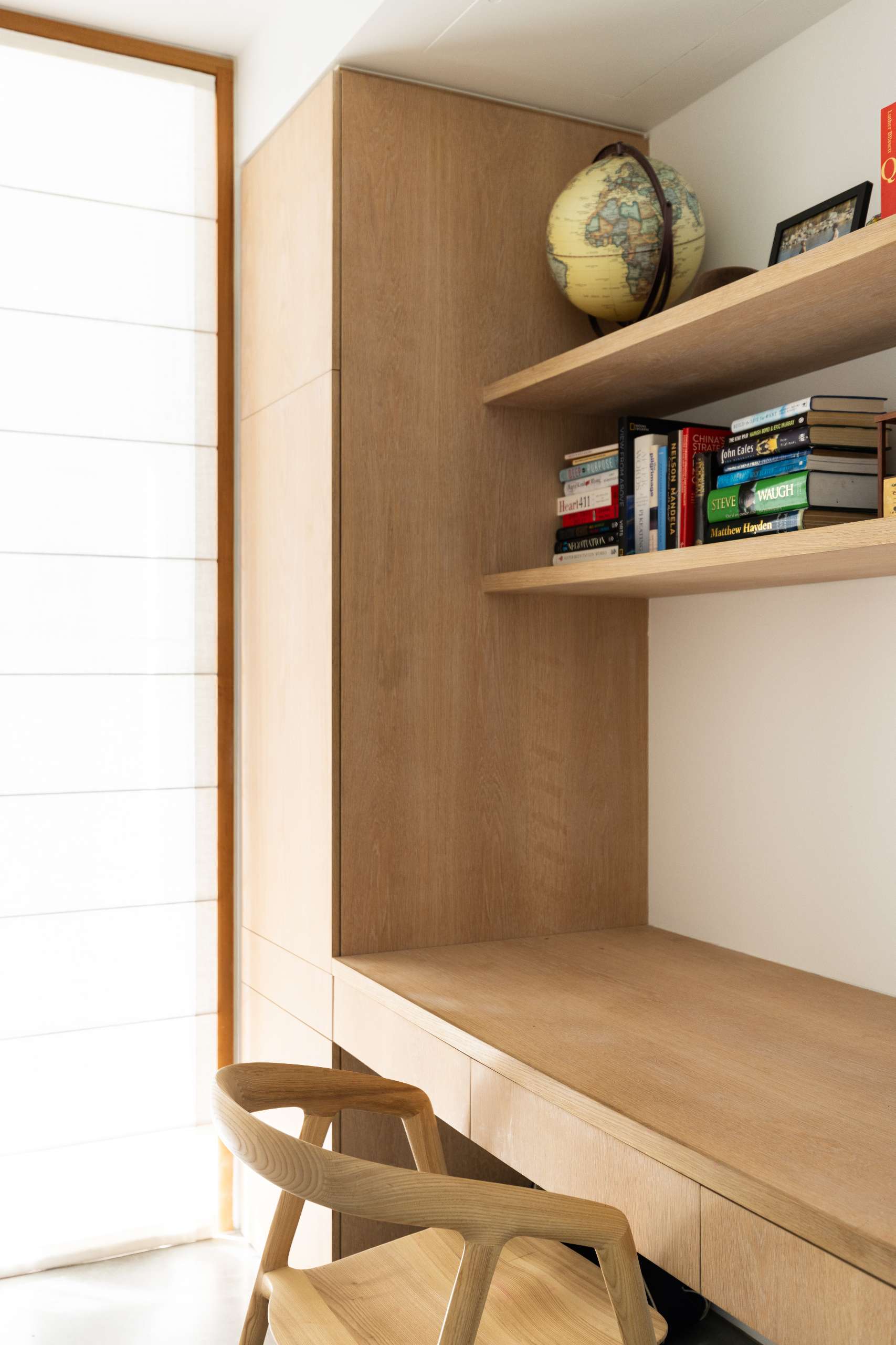 This study nook features custom joinery in warm wood, with integrated shelves and a built-in desk that maximizes space. A timber chair complements the natural palette, while filtered light from the sheer Roman blind enhances the cozy, focused atmosphere.