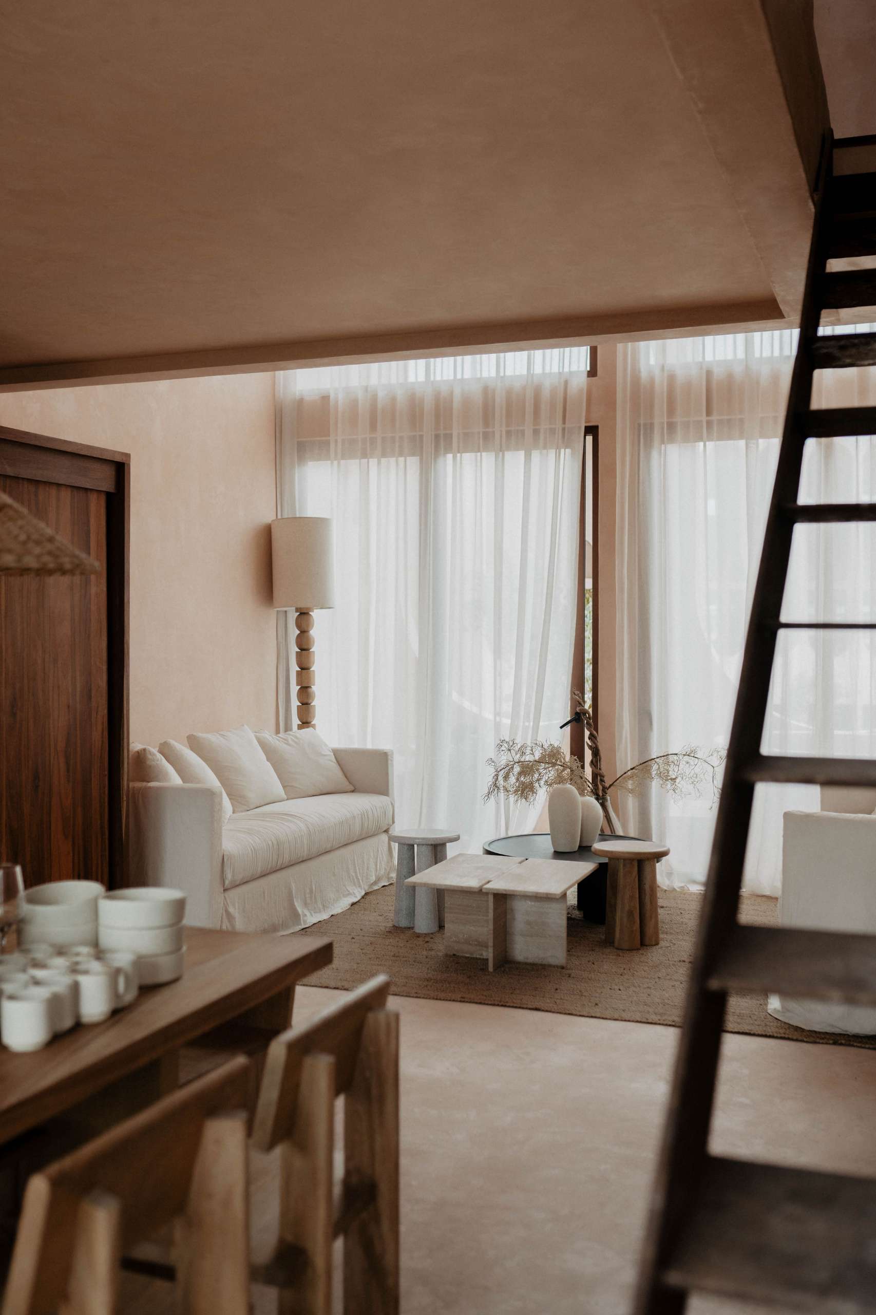 In this apartment, neutral tones and organic materials take center stage in the living room. Filtered sunlight washes over a minimalist sofa, sculptural wood tables, and a layered jute rug, creating a space that feels both serene and elemental.