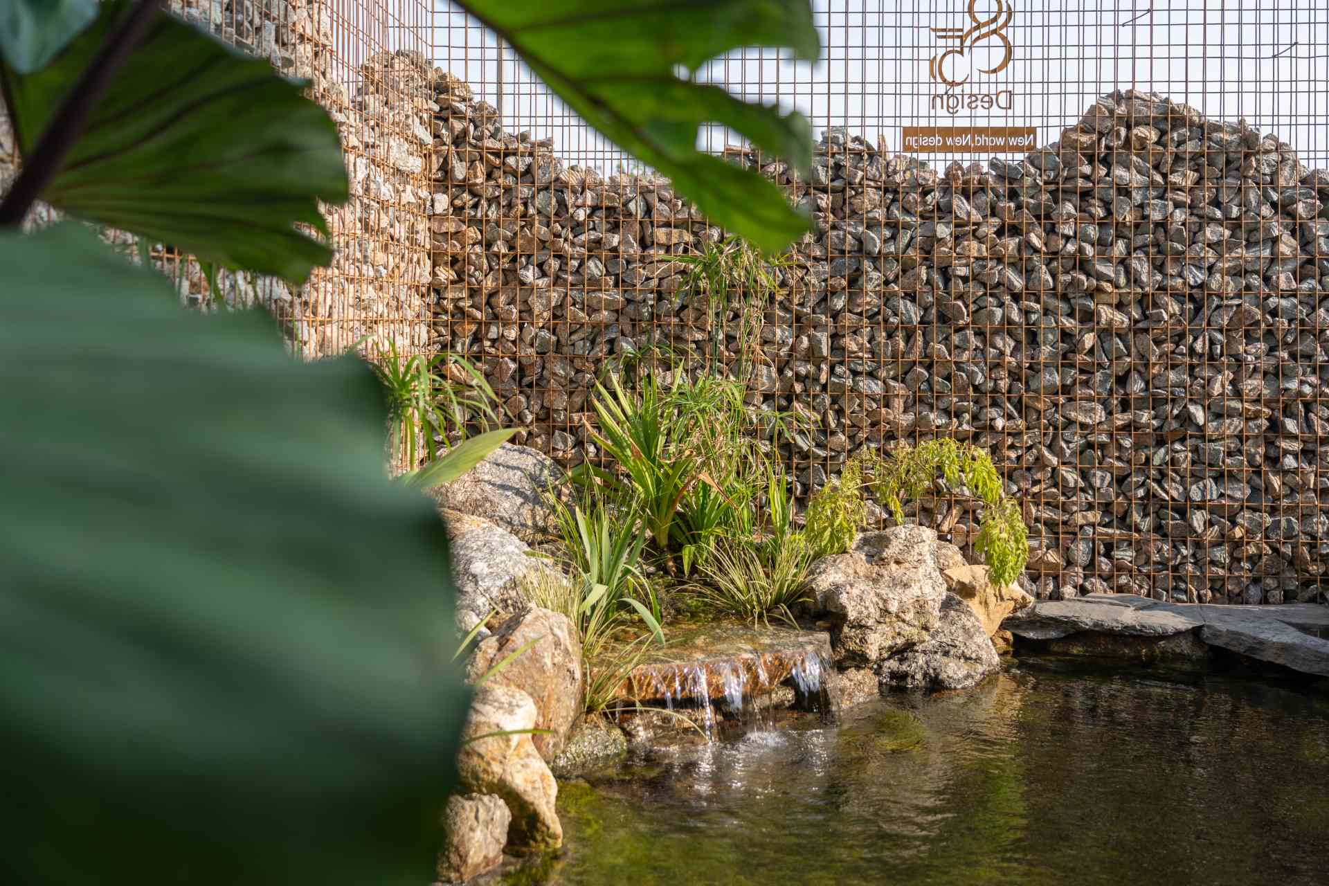 A glass volume suspended above a pond creates a striking waterfall that cools the space and adds a calming sound. Surrounded by tropical plants, natural stone, and a textured gabion wall, the garden feels like a small, living ecosystem built into the heart of a modern home.
