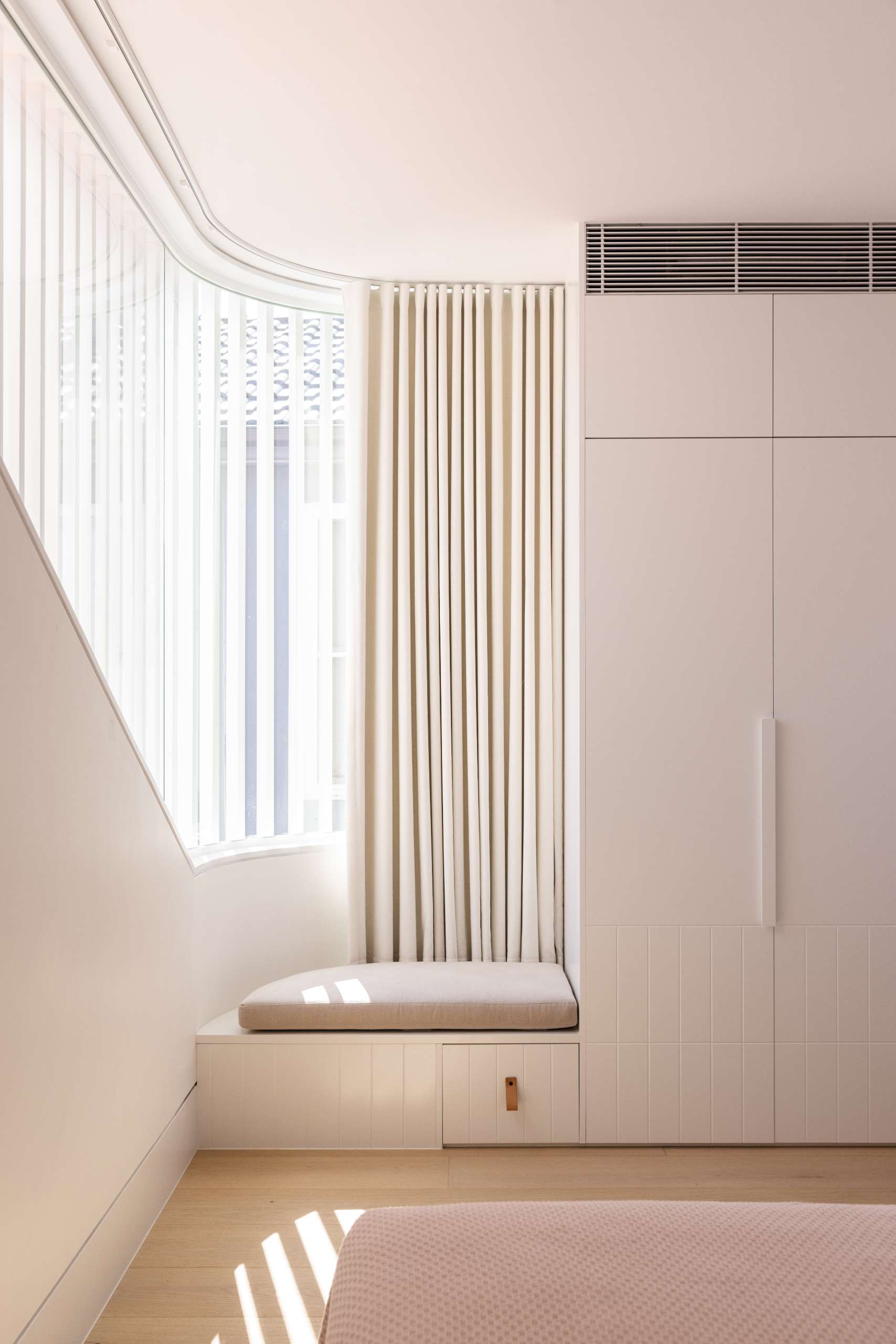 In this bedroom, there's a corner featuring a softly curved window, while a custom-built window seat with integrated storage below adds functionality and a cozy spot to rest. The pale timber floor and tonal cabinetry maintain a calm, cohesive palette.