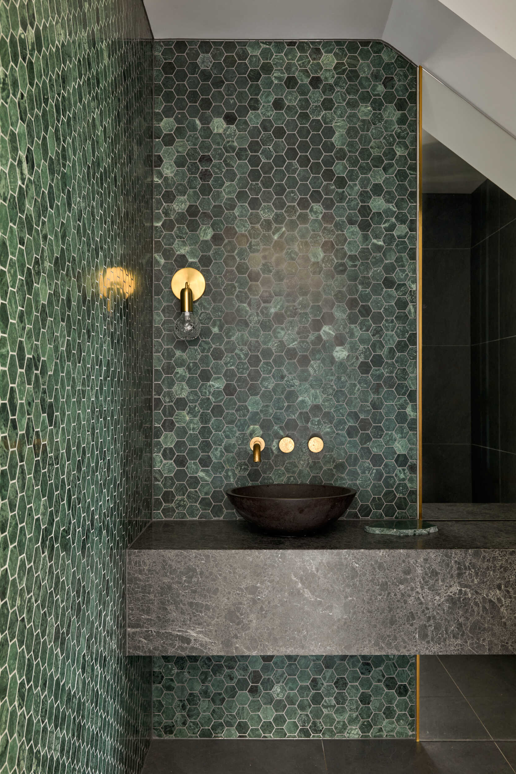 The powder room makes a confident statement, wrapping its walls in a sea of emerald hexagonal tiles that shimmer with depth and texture. The green tones shift gently between moss and jade, giving the surface a natural variation that feels alive and richly layered. Against this vibrant backdrop, the matte black vessel sink sits like a smooth river stone, grounded by the solid heft of the gray stone vanity. Brass fixtures add a dash of warmth and a hint of glamour, catching the light just enough to draw the eye without stealing the show.