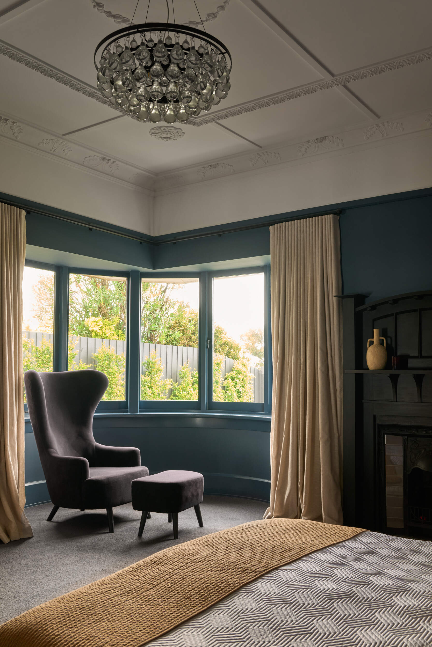 Moody blue walls wrap around this bedroom, grounding it with a sense of quiet elegance, while the soft daylight filters through the bay window and gently brightens the room. The ceiling’s original plaster detailing brings a touch of vintage charm, echoed by the shimmering glass teardrops of the chandelier above. Meanwhile, a dark fireplace mantle adds weight and heritage, styled with just a few simple ceramic vessels for an understated finish.