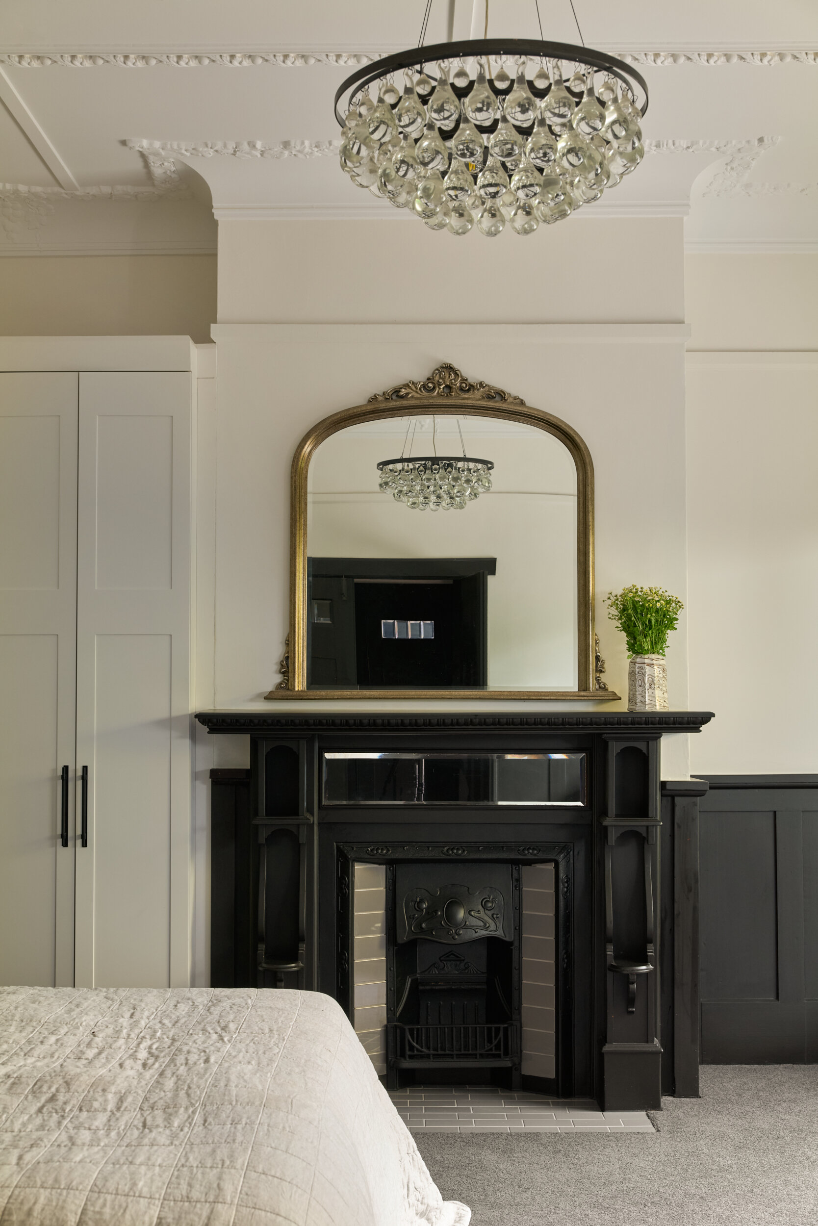 This bedroom is a graceful blend of old-world charm and quiet sophistication. At its center, a classic black fireplace holds court with intricate molding and a finish that plays gently with the light. Above it, an antique-style mirror framed in soft gold reflects a teardrop chandelier, echoing the sparkle overhead and doubling the sense of height and airiness.