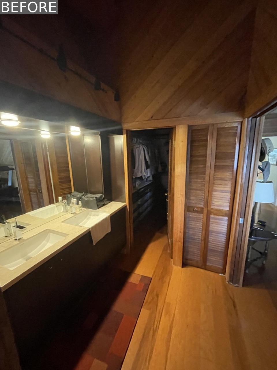 BEFORE - The original primary bathroom felt dark and enclosed, with heavy wood paneling extending across the walls and ceiling. A long double vanity with under-mounted sinks sat beneath a soffit of dated lighting, further dimming the space. The surrounding closet doors and cabinetry blended into the wood-heavy backdrop, creating a monotone, almost cabin-like feel that lacked contrast and visual relief.