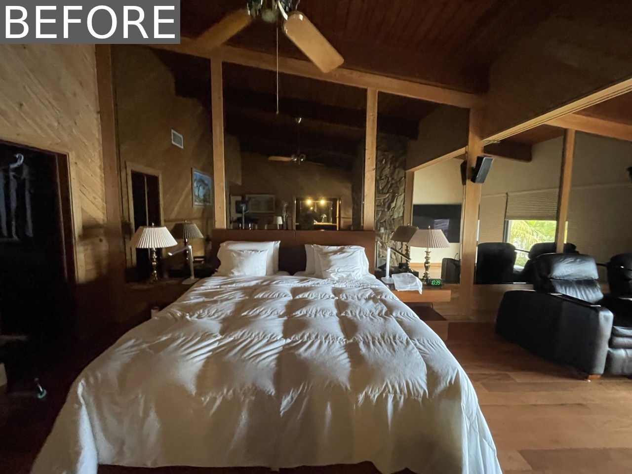 BEFORE - The original primary bedroom felt dark and dated, with a full mirrored wall, a heavy stone accent, and a pair of oversized black leather recliners focused on a TV.