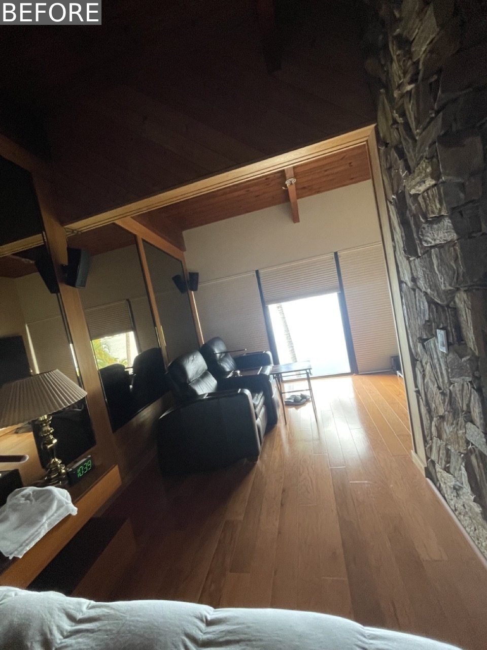 BEFORE - The original primary bedroom felt dark and dated, with a full mirrored wall, a heavy stone accent, and a pair of oversized black leather recliners focused on a TV.