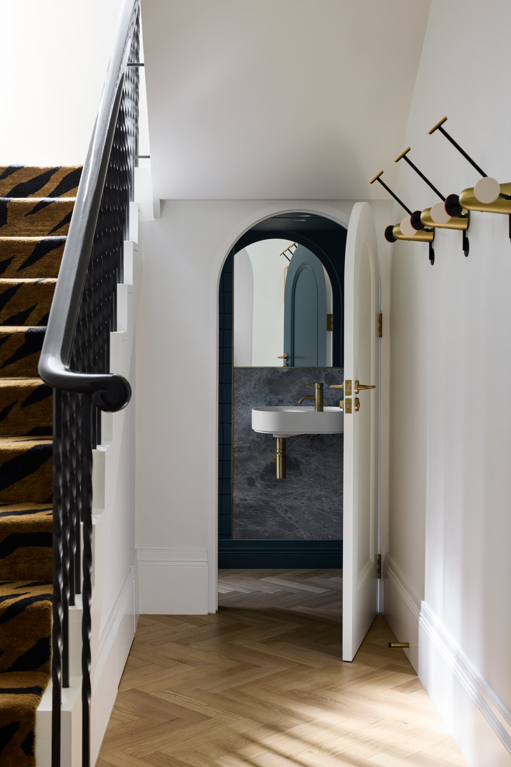 This powder room, though compact, reflects the same level of craft and material sensitivity as the rest of the house. The small room feels like a little design secret, revealed through a curved doorway, and framed in deep blue. The cool marble wall and brushed brass fixtures add polish, while the simple white basin keeps things feeling light and minimal.