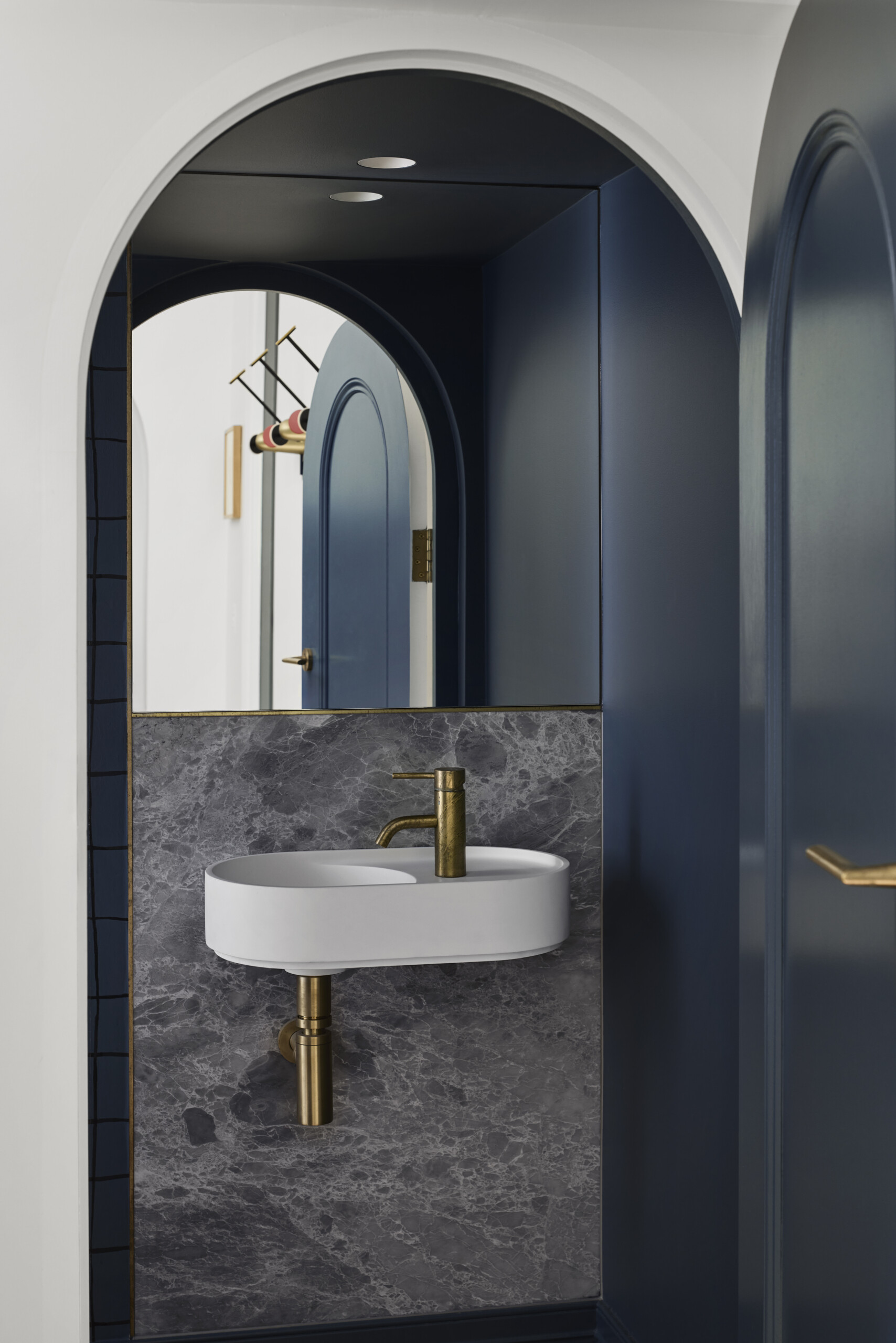 This powder room, though compact, reflects the same level of craft and material sensitivity as the rest of the house. The small room feels like a little design secret, revealed through a curved doorway, and framed in deep blue. The cool marble wall and brushed brass fixtures add polish, while the simple white basin keeps things feeling light and minimal.