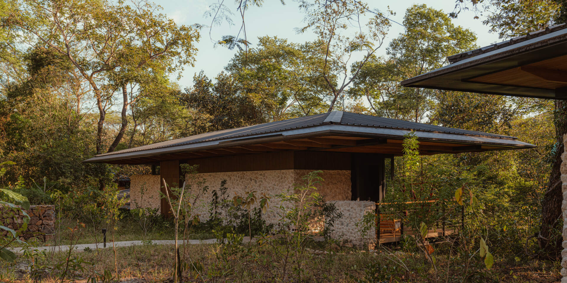 A stone-clad jungle bungalow in Costa Rica blends effortlessly into its forest surroundings. A curved concrete path leads to the entrance, while a floating terrace and rich timber ceiling invite guests into a space that feels both grounded and elevated.