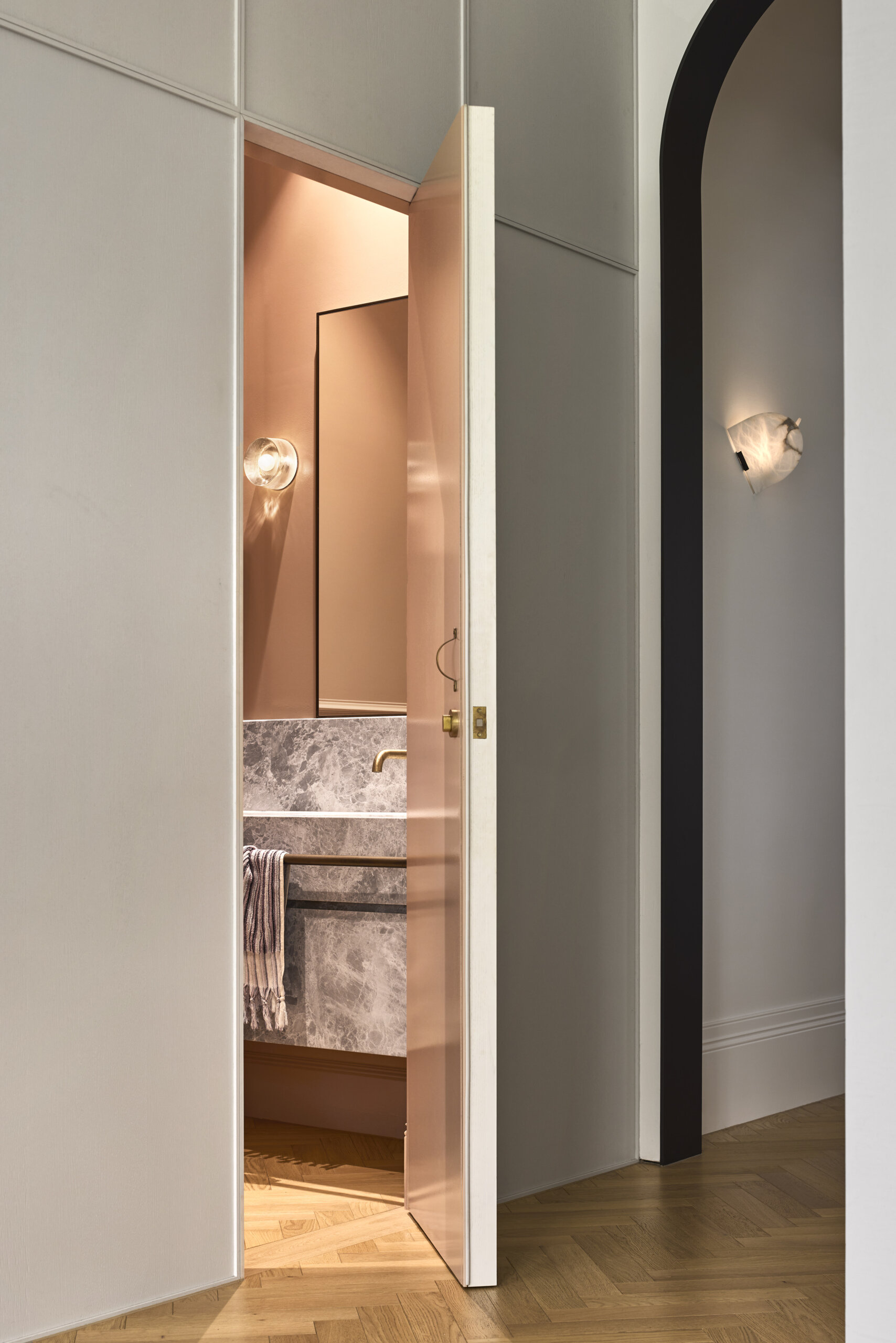Partially revealed through a slim, glossy door, this bathroom glows with quiet drama. The warm blush walls and matching cabinetry create a cocoon-like feel, while soft lighting and polished brass accents give the space a gentle elegance. Cool gray marble adds texture and contrast beneath the sink, anchoring the palette with natural depth.