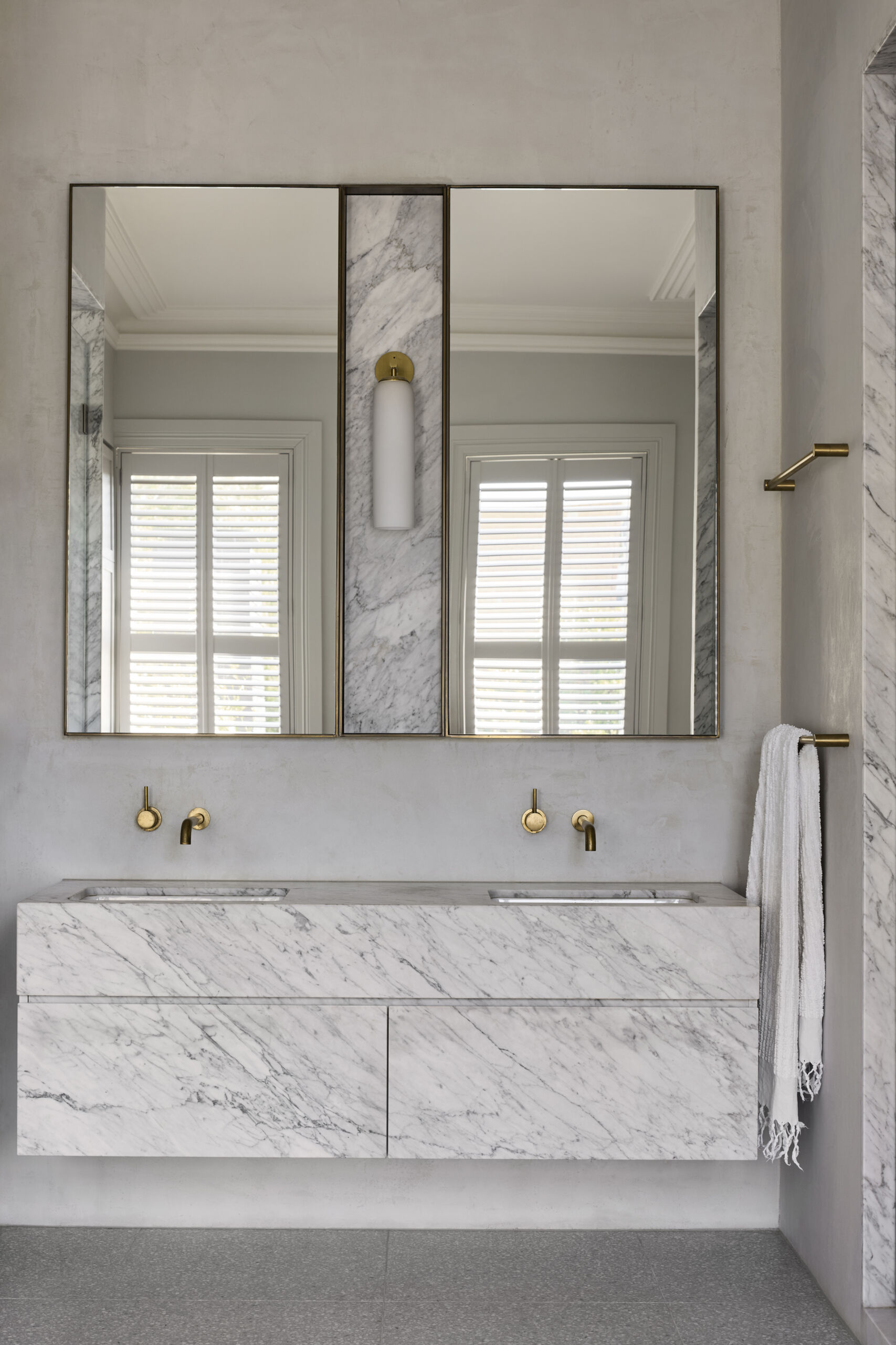 This bathroom is has a double vanity that's all about quiet confidence, wrapped in seamless marble that shows off its natural veining like brushstrokes on a canvas. Brass fixtures punctuate walls with warmth and elegance, while the mirrors are split by a simple vertical sconce that echoes the symmetry of the whole design.