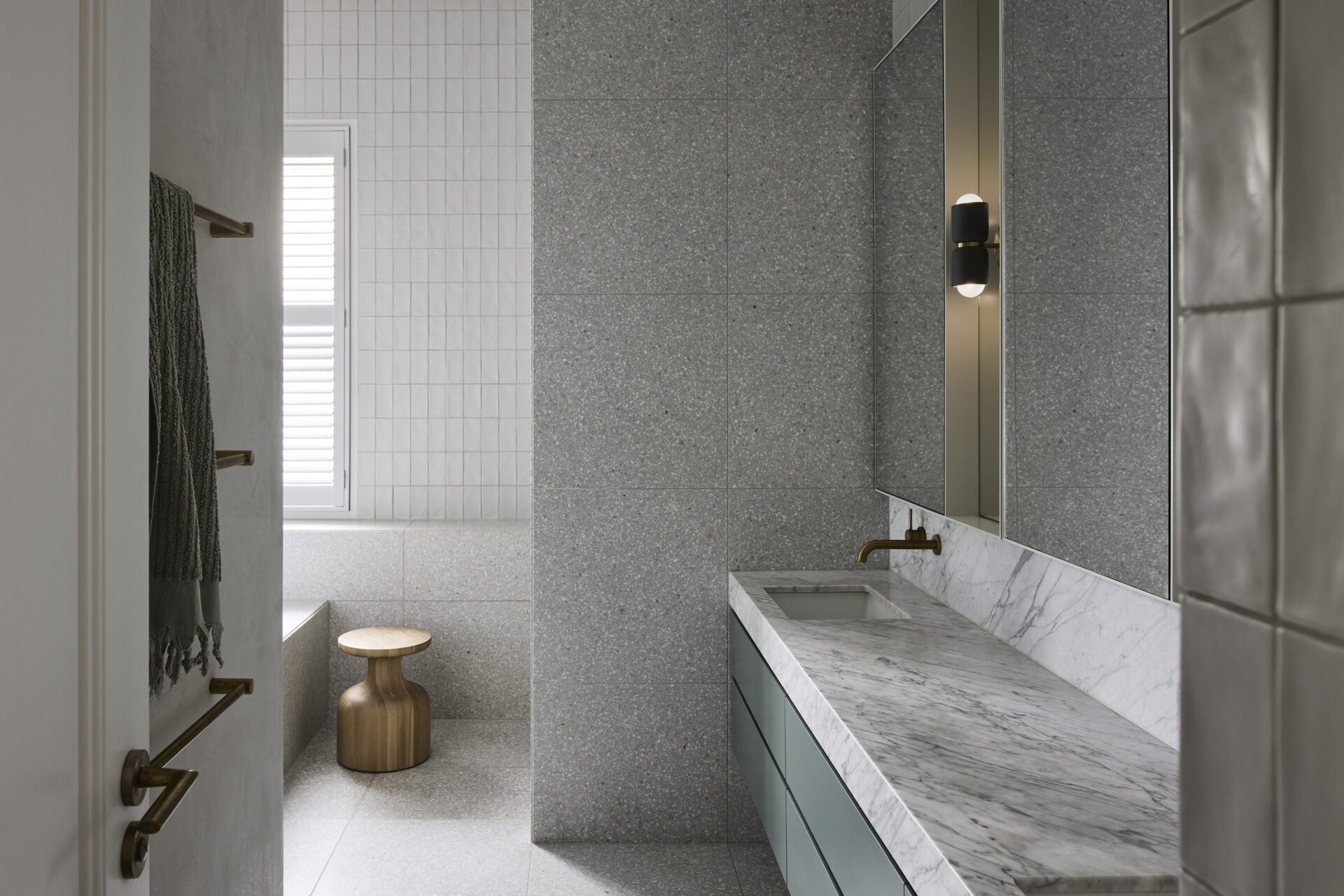 This bathroom includes muted sage vanity floating beneath a long marble countertop, where soft veining introduces a subtle rhythm. Walls are lined in pale terrazzo and hand-finished tiles, offering a balance of crisp geometry and warmth. Brass tapware and black sconces provide contrast without drawing focus.