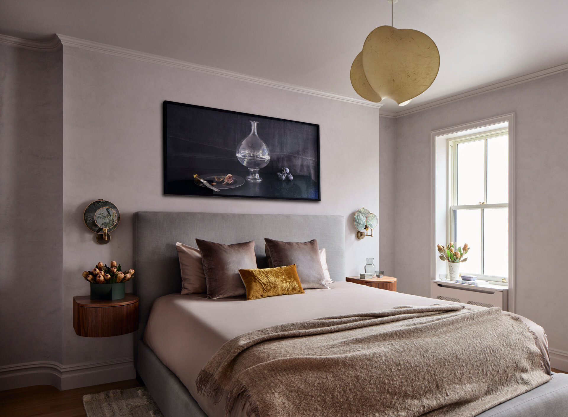 This primary suite is softened with plaster in a lavender-gray hue, complemented by stained oak elements. Custom floating nightstands are lit by printed linen sconces, and a vintage resin pendant hangs overhead. A framed still life adds a final personal touch.
