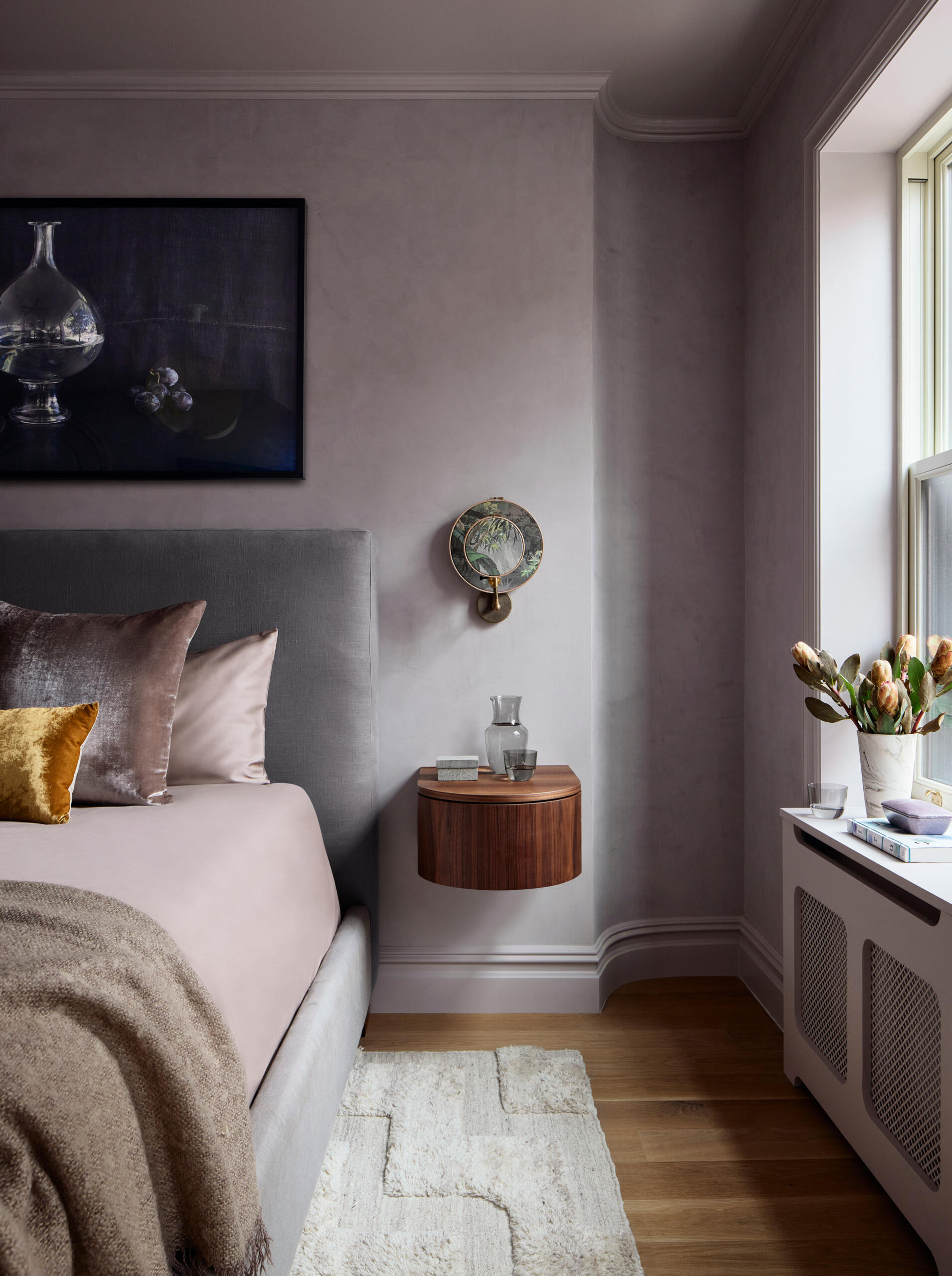 This primary suite is softened with plaster in a lavender-gray hue, complemented by stained oak elements. Custom floating nightstands are lit by printed linen sconces, and a vintage resin pendant hangs overhead. A framed still life adds a final personal touch.