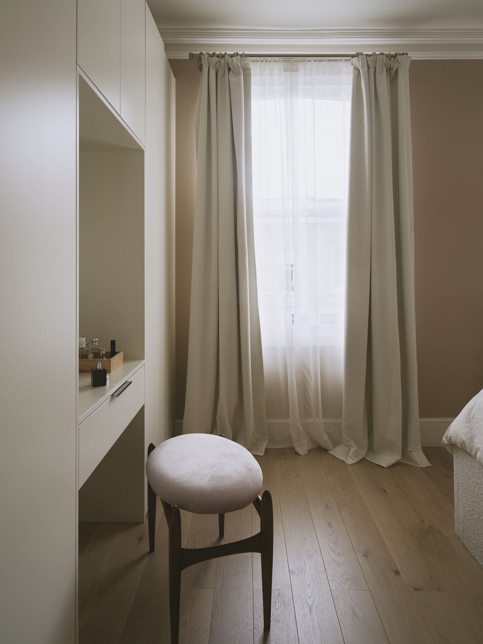 Soft light filters through sheer curtains, giving this bedroom nook a quiet, dreamy atmosphere. The walls are painted in a muted tone that feels warm without overpowering, pairing beautifully with the wood flooring that adds subtle texture underfoot. A simple built-in vanity is tucked neatly against the wall, its matte white finish blending seamlessly with the cabinetry, while a low, rounded stool upholstered in pale velvet provides a seating for the vanity.