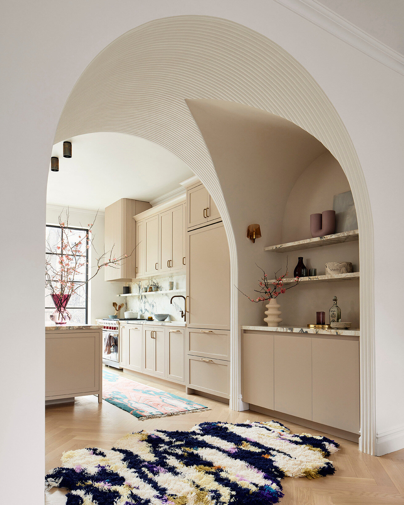 A deep fluted plaster arch creates a dramatic architectural gesture, guiding the eye from the living area toward the kitchen and rear garden beyond. Nestled within the arch, a discreet bar area is softly lit by vintage French smoked glass sconces, casting a warm, ambient glow.