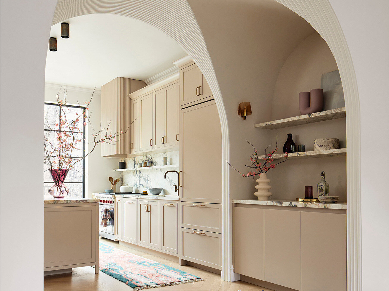 A deep fluted plaster arch creates a dramatic architectural gesture, guiding the eye from the living area toward the kitchen and rear garden beyond. Nestled within the arch, a discreet bar area is softly lit by vintage French smoked glass sconces, casting a warm, ambient glow.