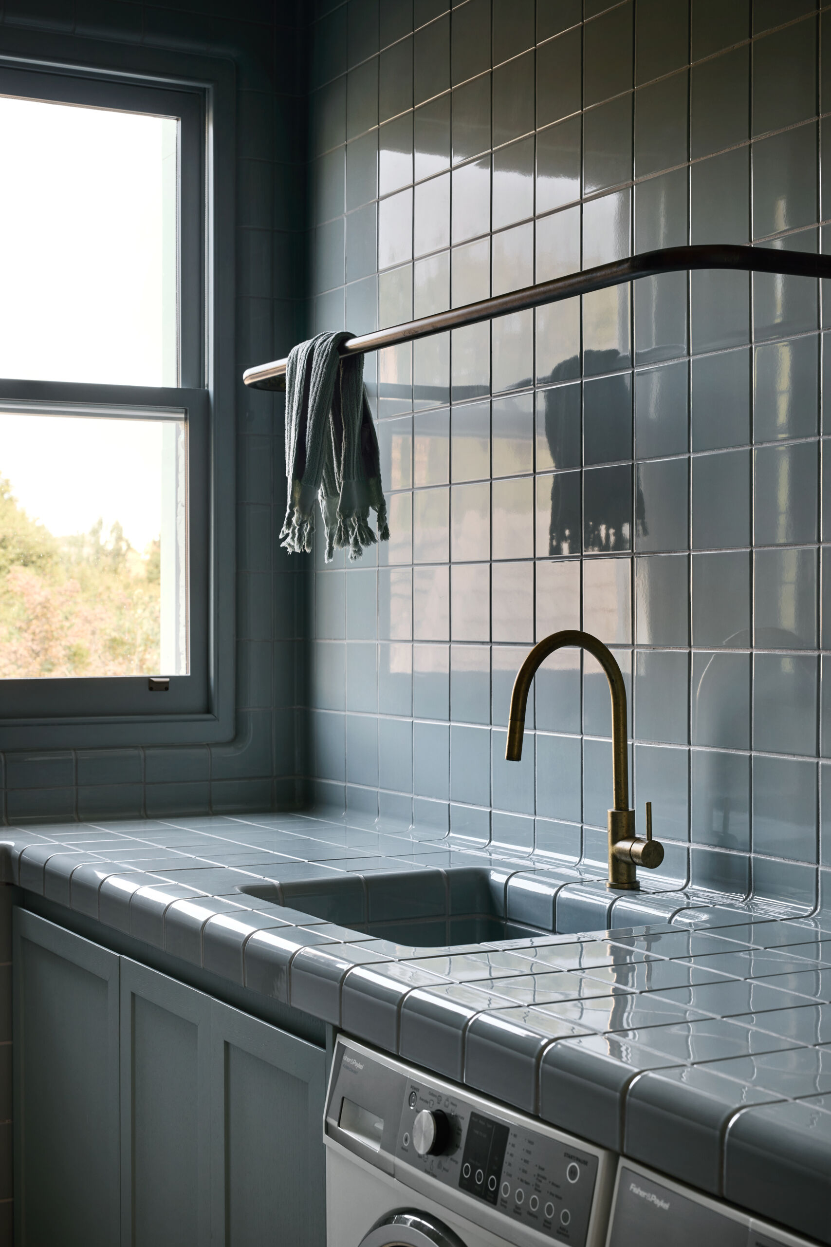With its soft blue tile and rounded edges, this laundry area feels unexpectedly serene and sculptural. The seamless transition from counter to sink adds a sense of flow, while the brass faucet introduces just the right amount of contrast and warmth. A single striped towel draped over a curved metal rail adds a relaxed, lived-in touch. Even the appliances tuck in quietly beneath the counter, letting the materials and textures take center stage. It’s a utility space that feels both functional and beautifully composed.