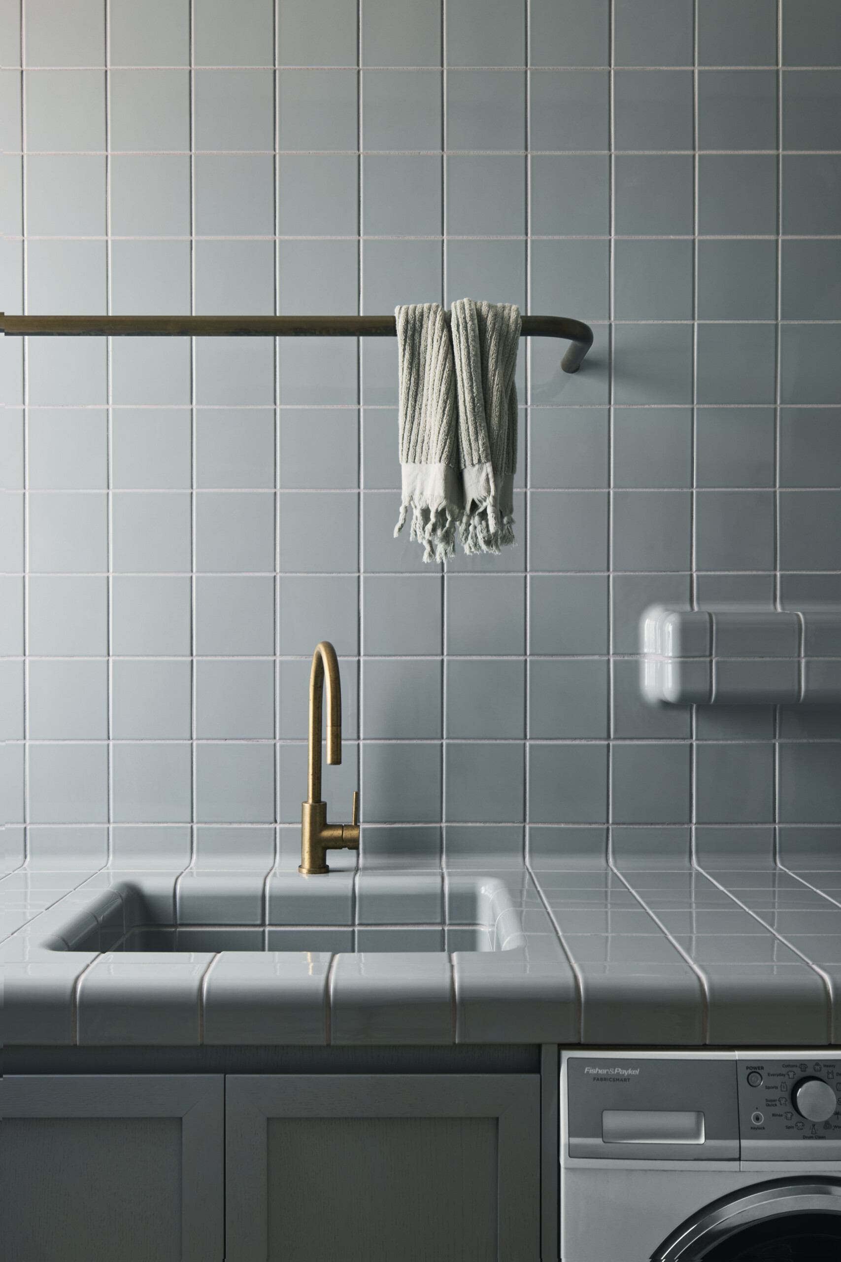 With its soft blue tile and rounded edges, this laundry area feels unexpectedly serene and sculptural. The seamless transition from counter to sink adds a sense of flow, while the brass faucet introduces just the right amount of contrast and warmth. A single striped towel draped over a curved metal rail adds a relaxed, lived-in touch. Even the appliances tuck in quietly beneath the counter, letting the materials and textures take center stage. It’s a utility space that feels both functional and beautifully composed.