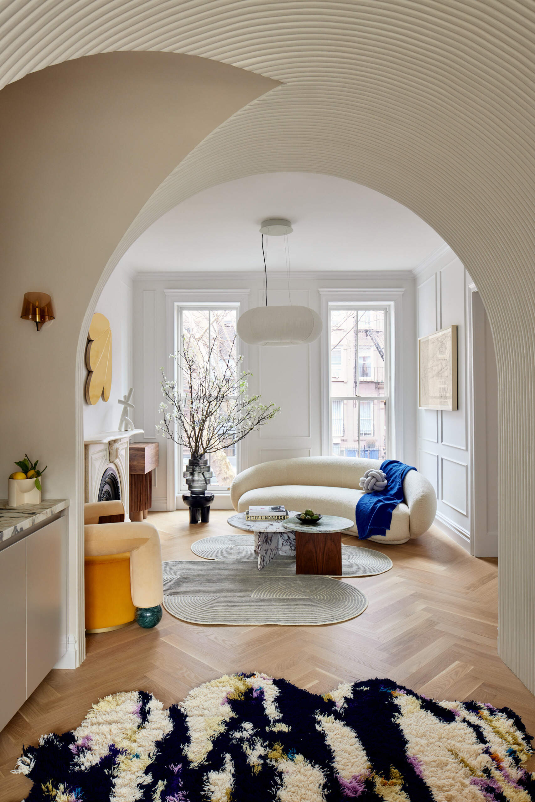 In the parlor, a curved sofa and matching armchair embrace two nested marble tables atop a fluidly shaped rug. A walnut sideboard anchors the space, while a gold-painted relief by Andrew Zimmerman and Cloud Support by Mike Hansel add a bold sculptural presence above the original marble fireplace mantel.