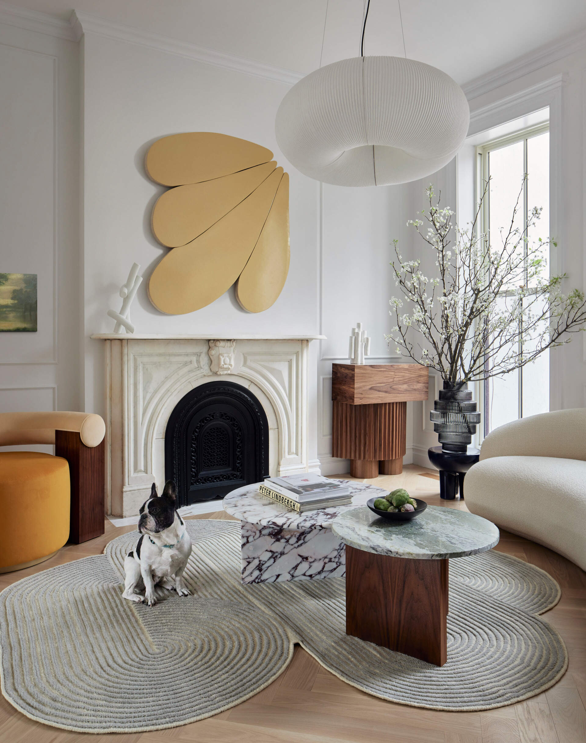In the parlor, a curved sofa and matching armchair embrace two nested marble tables atop a fluidly shaped rug. A walnut sideboard anchors the space, while a gold-painted relief by Andrew Zimmerman and Cloud Support by Mike Hansel add a bold sculptural presence above the original marble fireplace mantel.