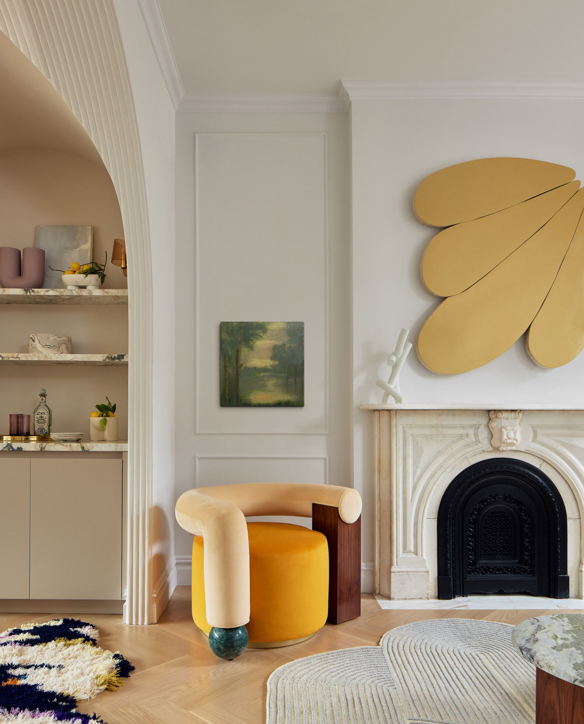 In the parlor, a curved sofa and matching armchair embrace two nested marble tables atop a fluidly shaped rug. A walnut sideboard anchors the space, while a gold-painted relief by Andrew Zimmerman and Cloud Support by Mike Hansel add a bold sculptural presence above the original marble fireplace mantel.