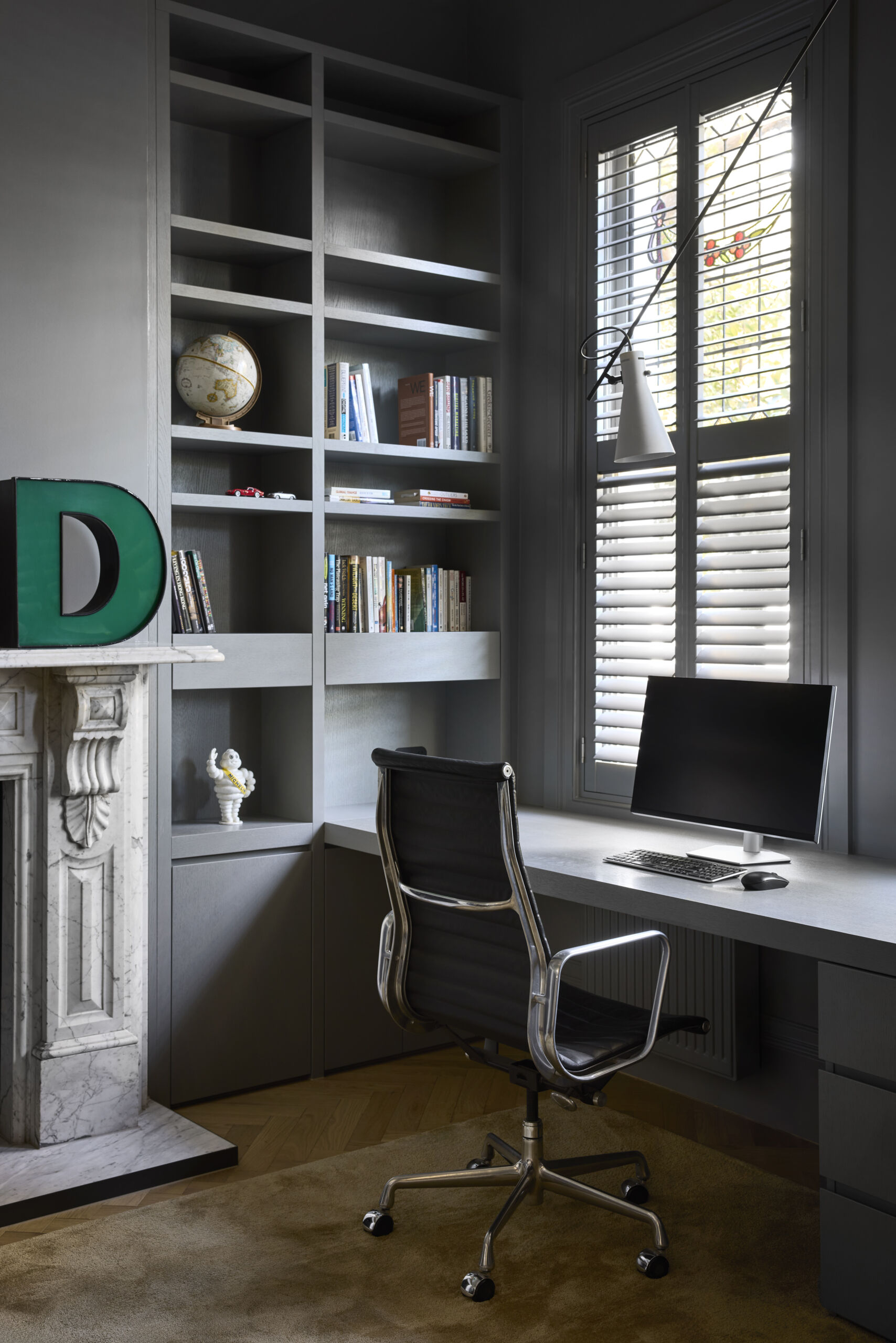 Moody and refined, the home office balances tradition and modern utility with ease. The rich gray cabinetry and built-in shelving give the room a tailored, grounded feel, while the sculptural desk lamp and sleek chair keep it firmly in the present. Natural light filters through the shutters, catching bits of stained glass and softly illuminating the crisp stone fireplace and pale wood floors.